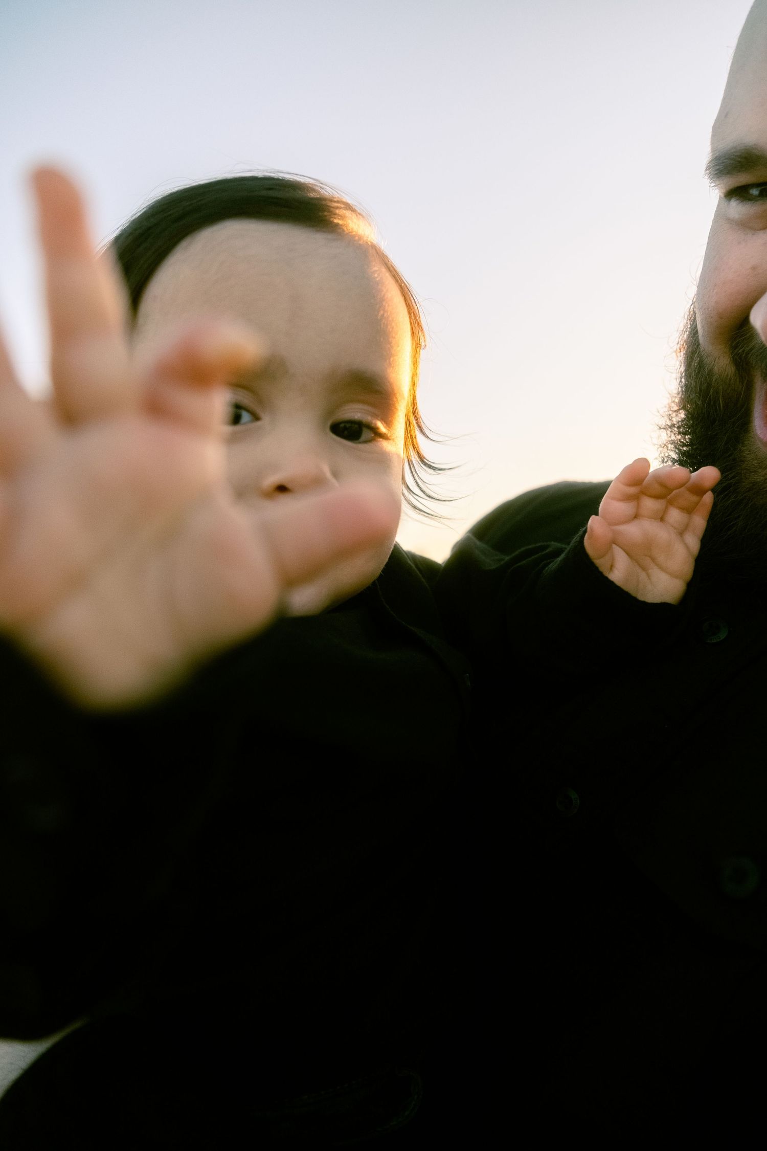 Bearded father holding baby in black outfit reaching toward camera at sunset outdoors.