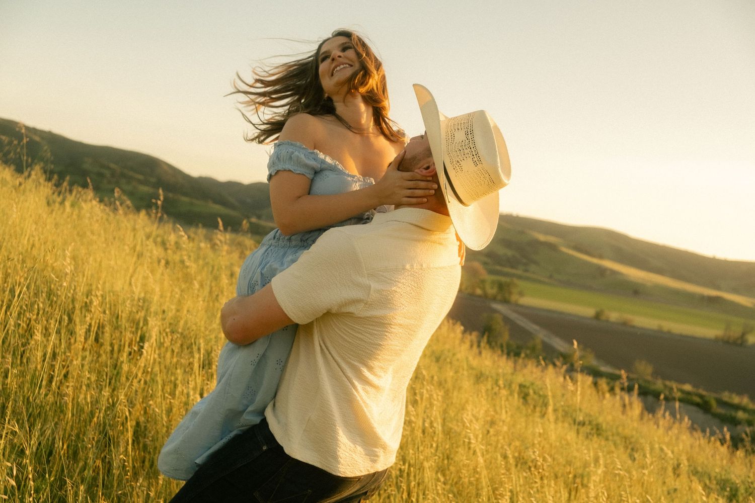 Man in white shirt and cowboy hat lifting woman in blue dress in golden sunlit hillside field.