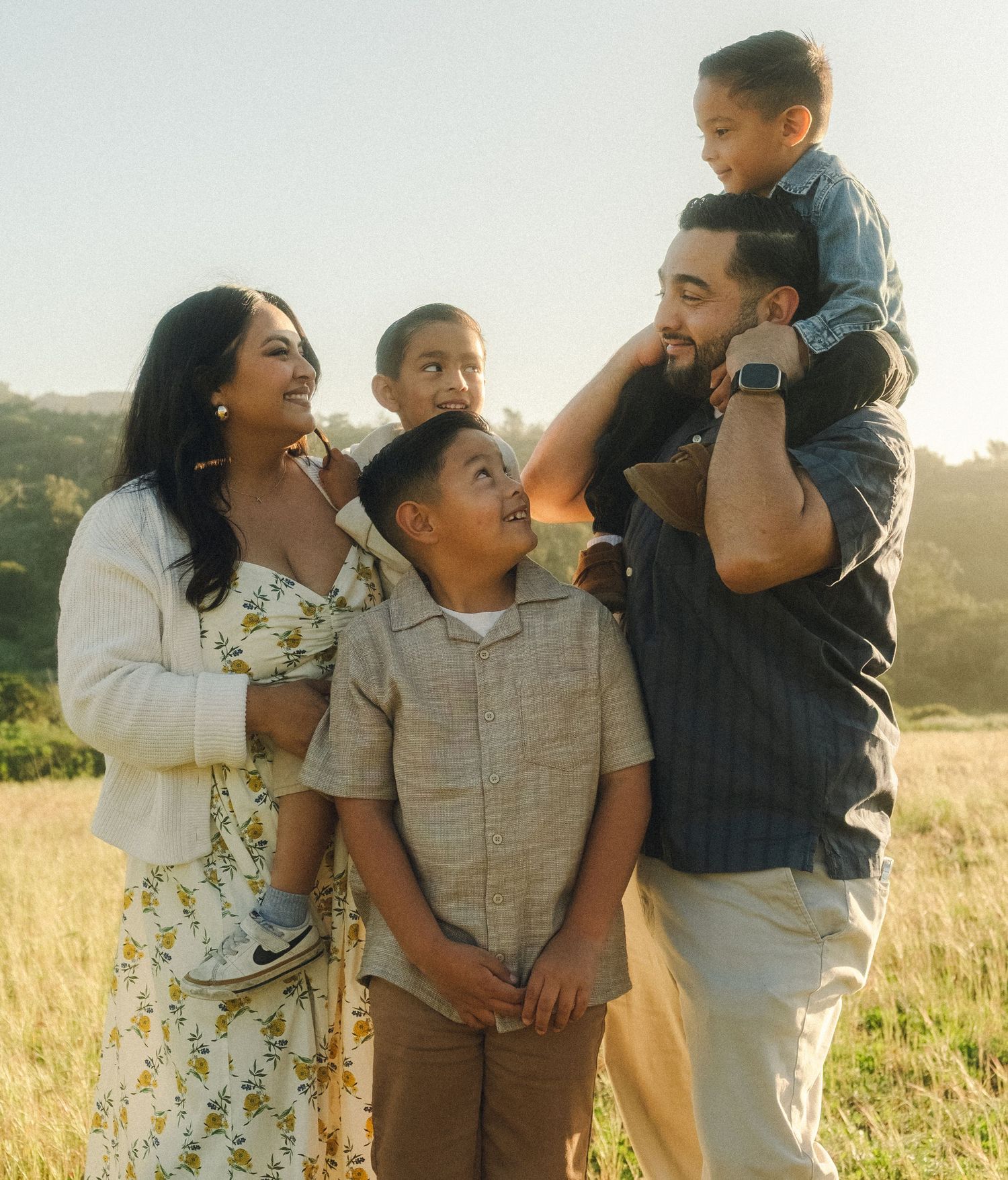 A joyful family of five poses outdoors in a sunny field, with parents and three children sharing smiles and laughter.