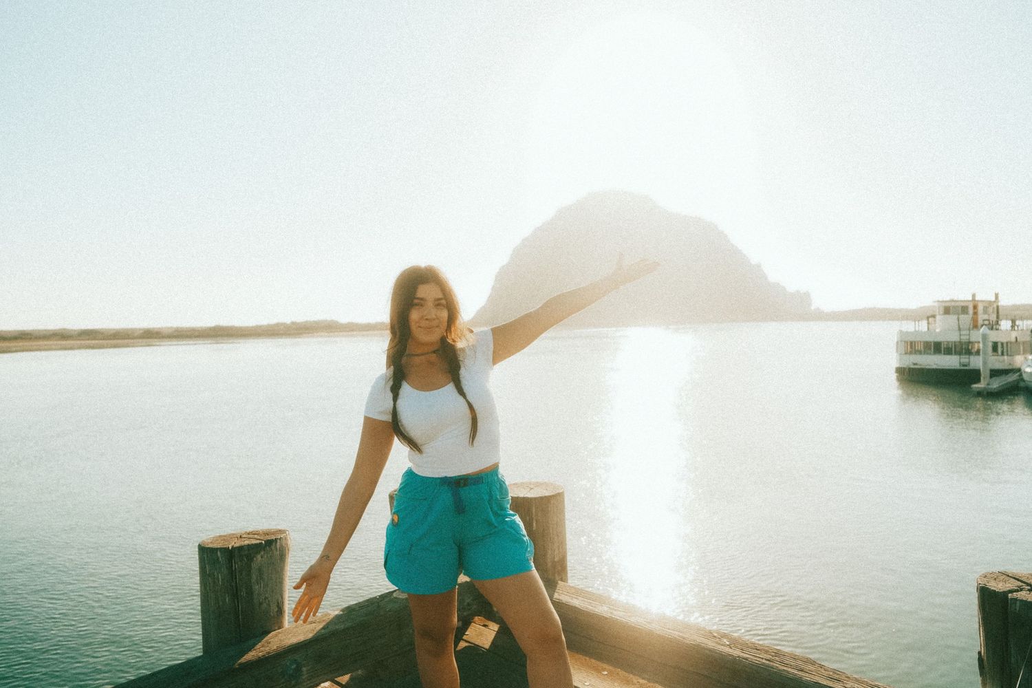 A person sits on a wooden dock overlooking a calm harbor at sunset with boats in the distance.
