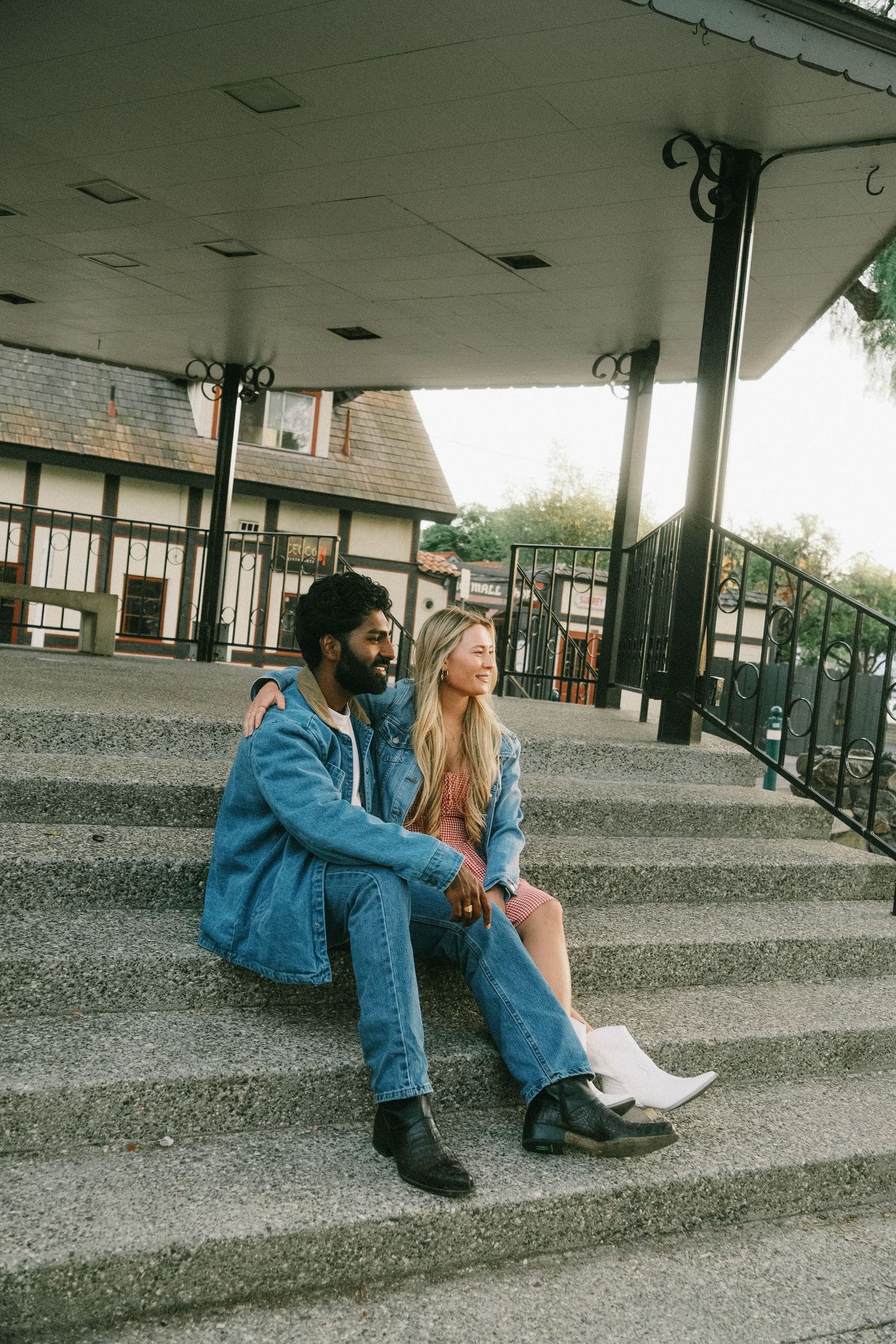A couple sits together on outdoor steps, both wearing denim outfits, sharing a tender moment near a building.