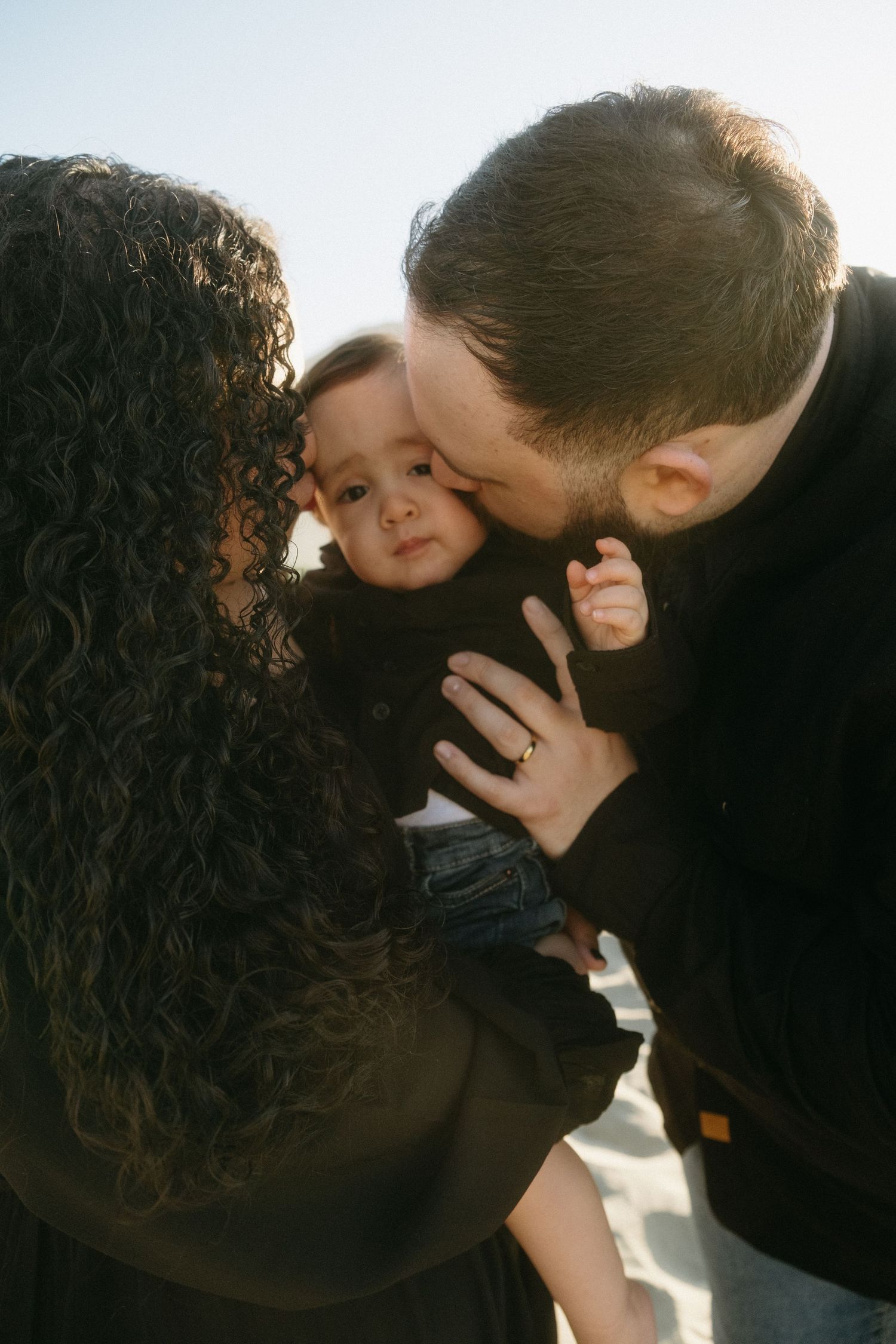 A couple lovingly kisses their baby outdoors, with the mother's curly hair and father's beard framing the moment.