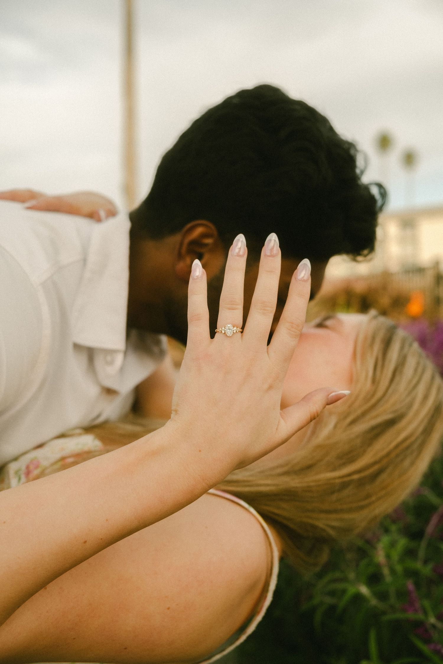 Couple sharing a romantic kiss outdoors, woman displaying diamond engagement ring with manicured nails in foreground.