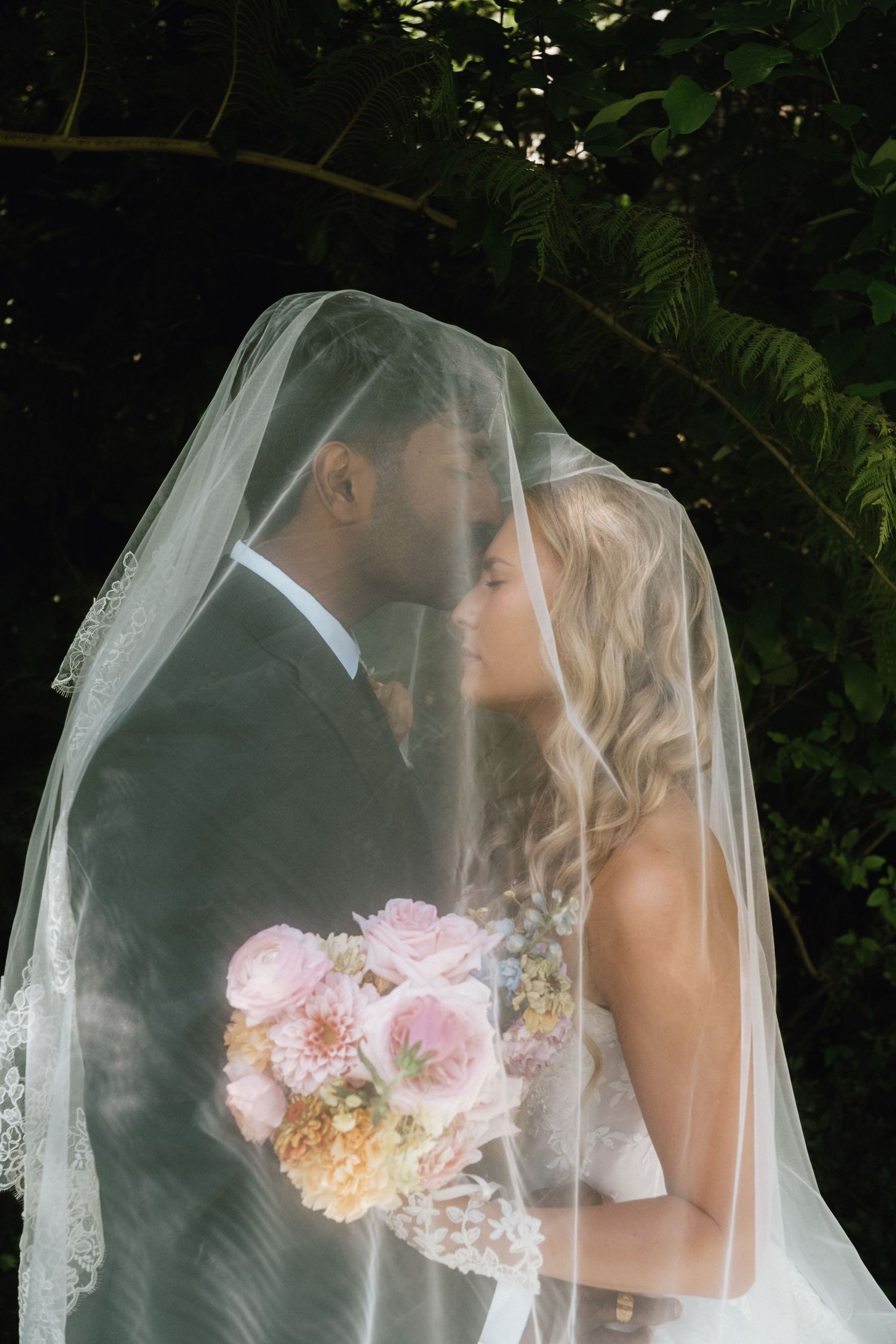 Bride and groom sharing a kiss under a sheer veil, bride holding a pink floral bouquet, surrounded by lush greenery.