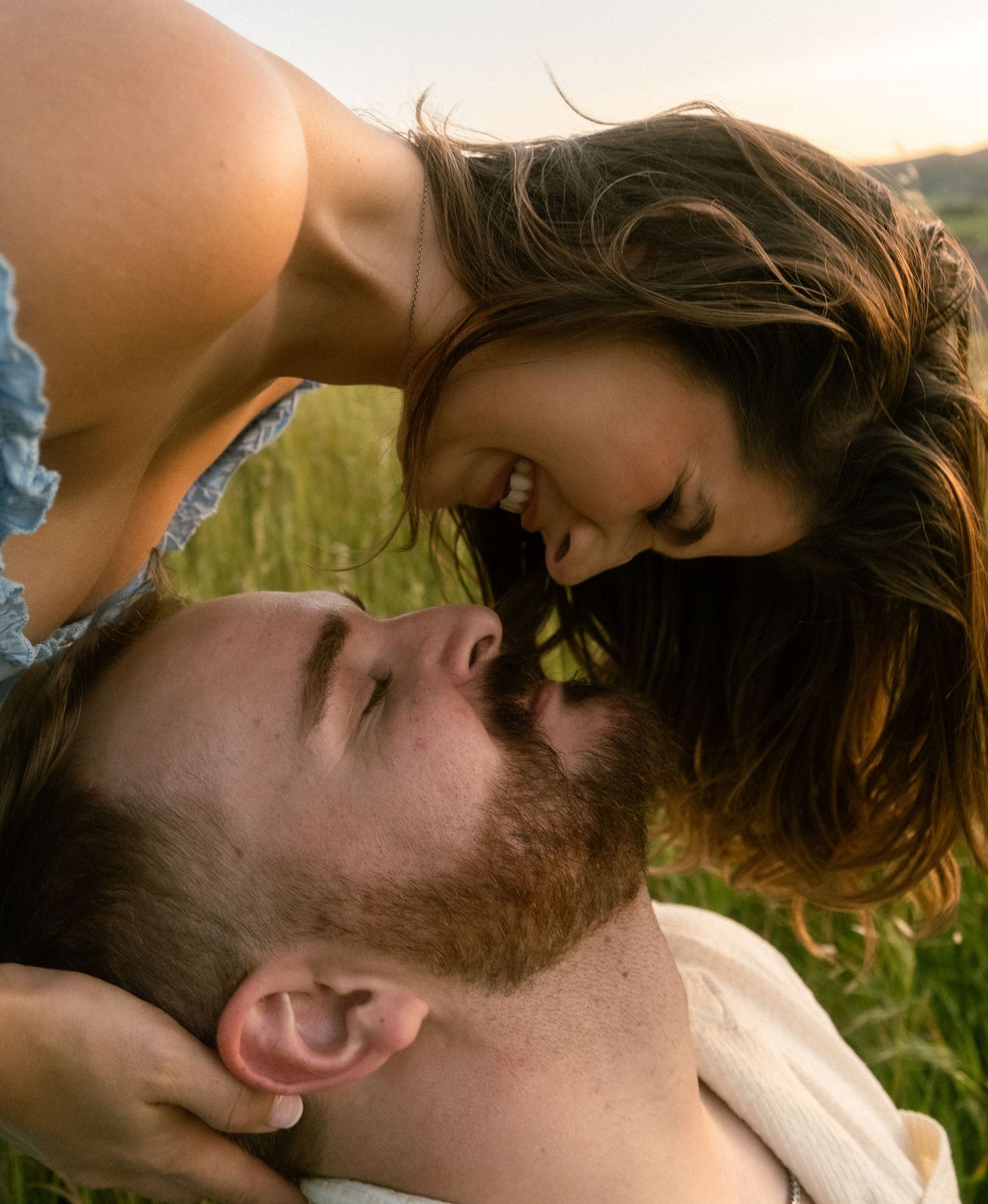 A couple shares a romantic moment outdoors, with a woman leaning over a man lying in a lush green field at sunset.
