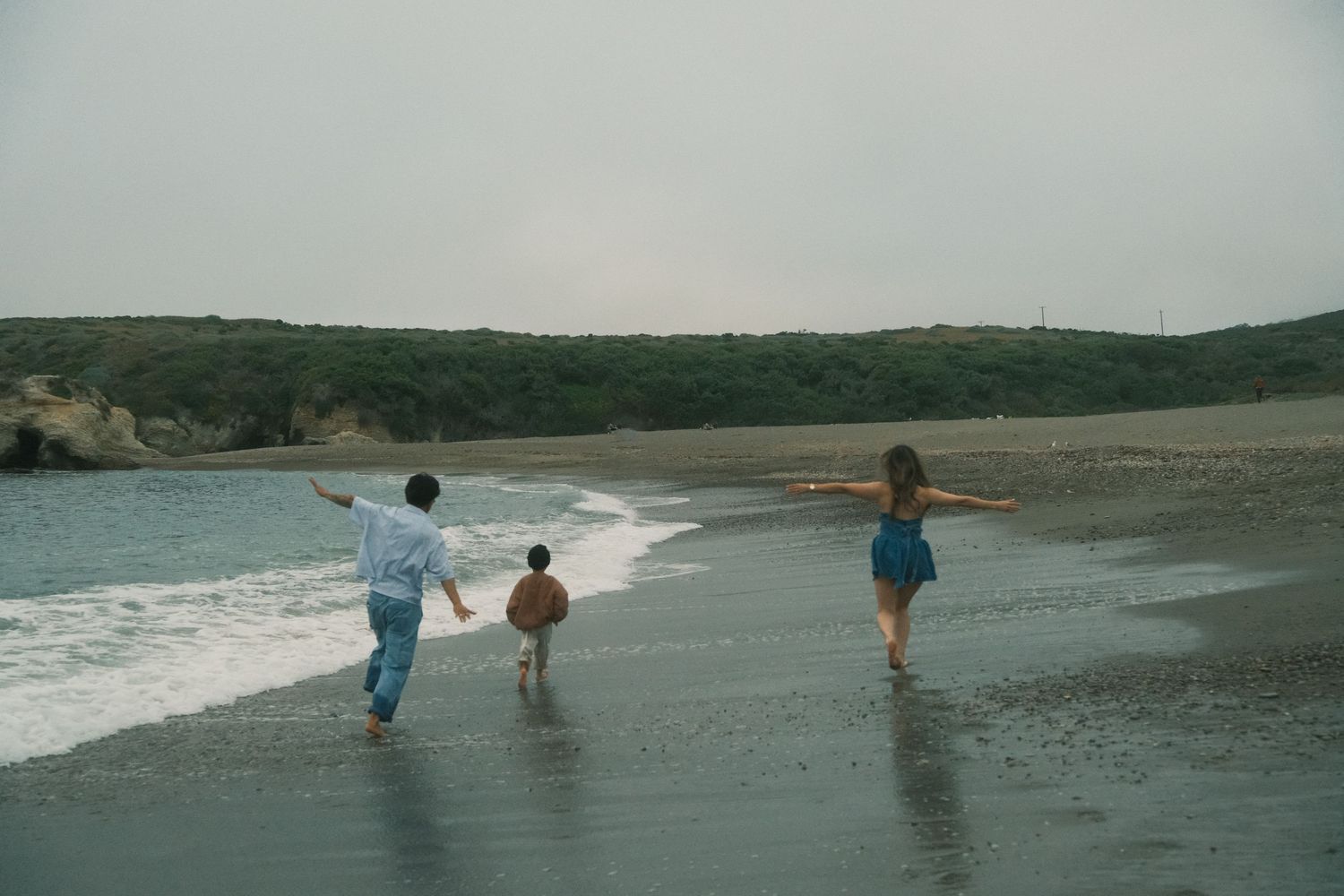 Three children running along a sandy beach shore with waves and green hills in the background on an overcast day.
