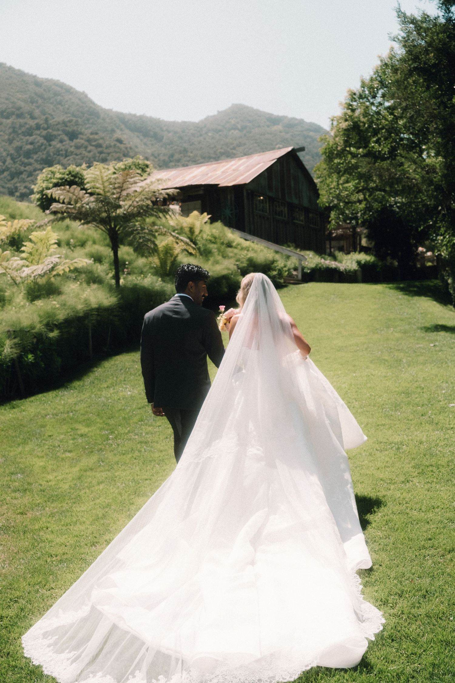 Bride and groom walking hand in hand on lush green lawn with rustic barn and mountain backdrop.