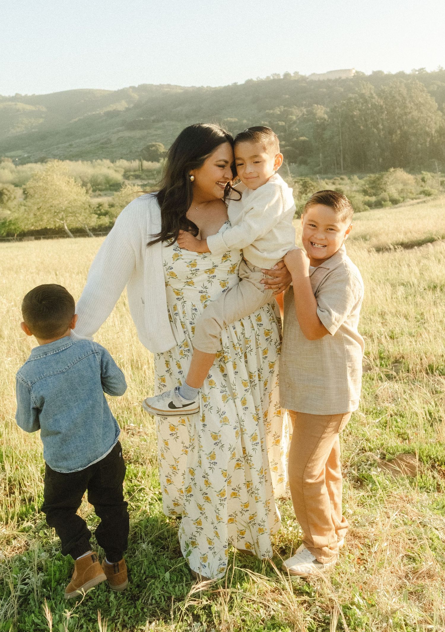A mother holds her toddler while two young boys smile outdoors in a sunny green field with hills in the background.