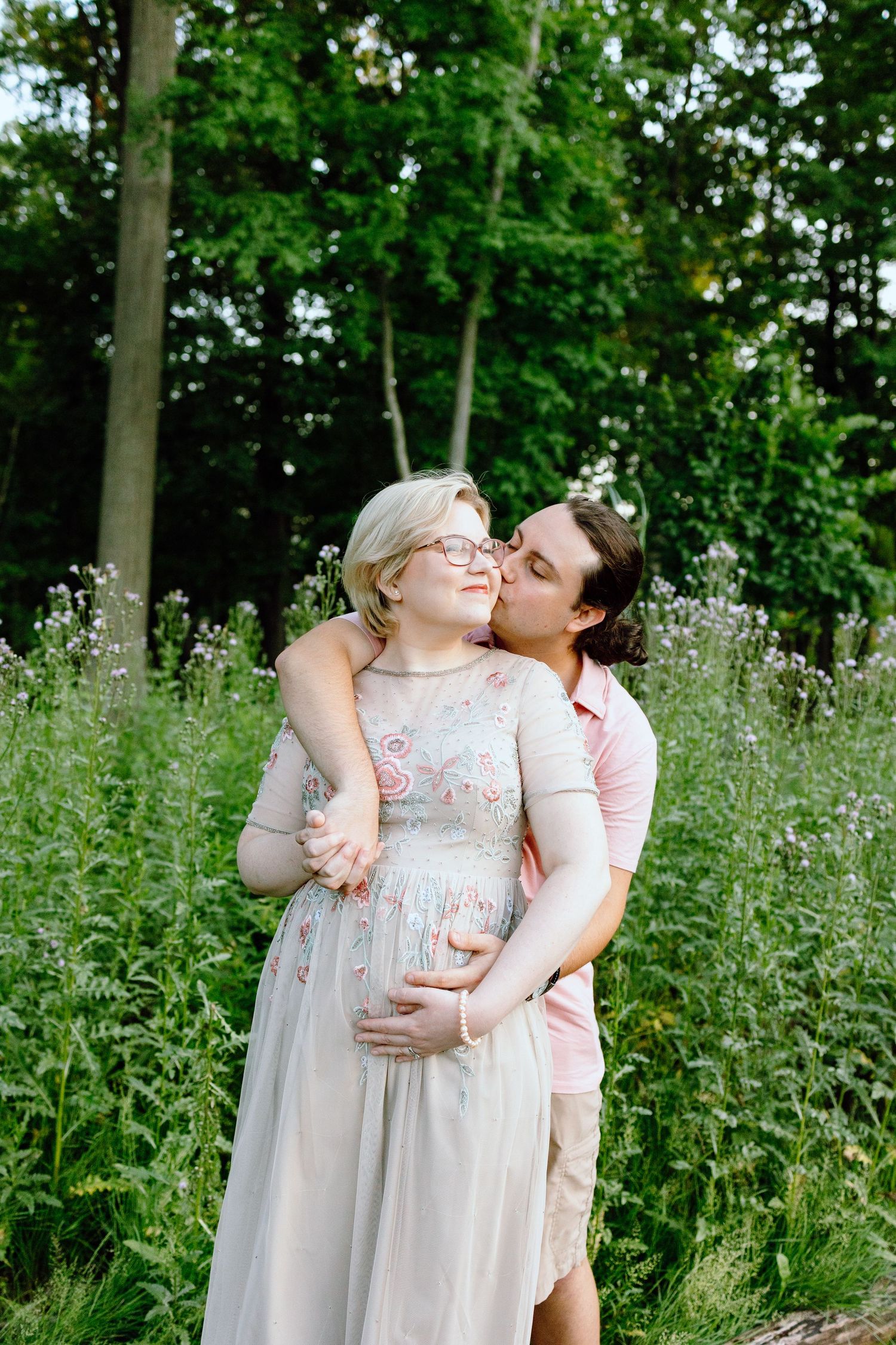 Husband kissing pregnant wife’s cheek in front of thistle bush in Beverly Hills Village Park, MI