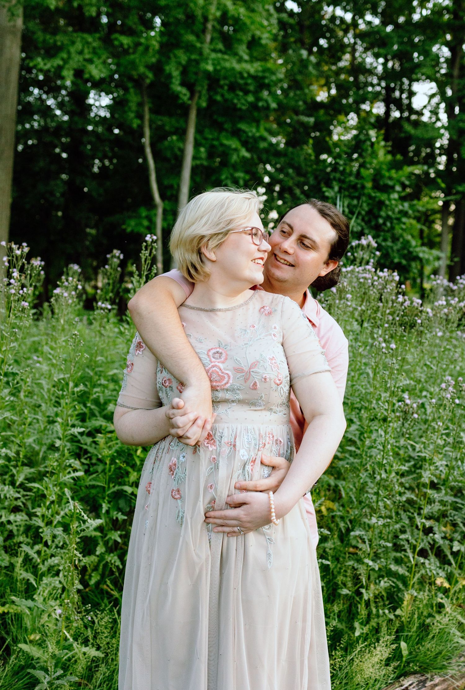 Pregnant couple embracing in front of thistle bush in Beverly Hills Village Park, MI