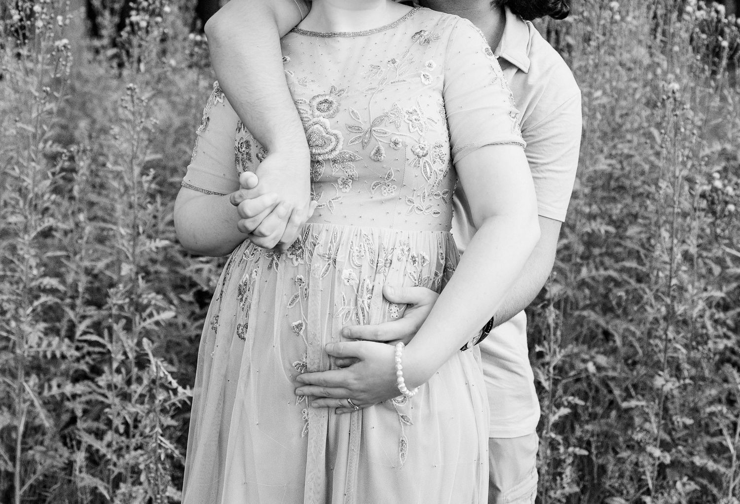 Pregnant couple embracing in front of thistle bush in Beverly Hills Village Park, MI