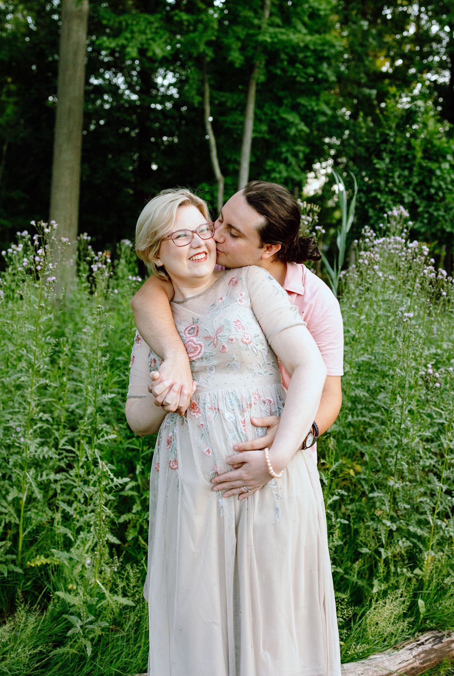 Husband kissing pregnant wife’s cheek in front of thistle bush in Beverly Hills Village Park, MI