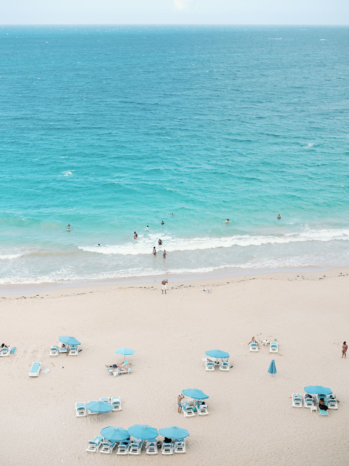 Aerial view of a turquoise beach with scattered blue umbrellas and lounge chairs on white sand.