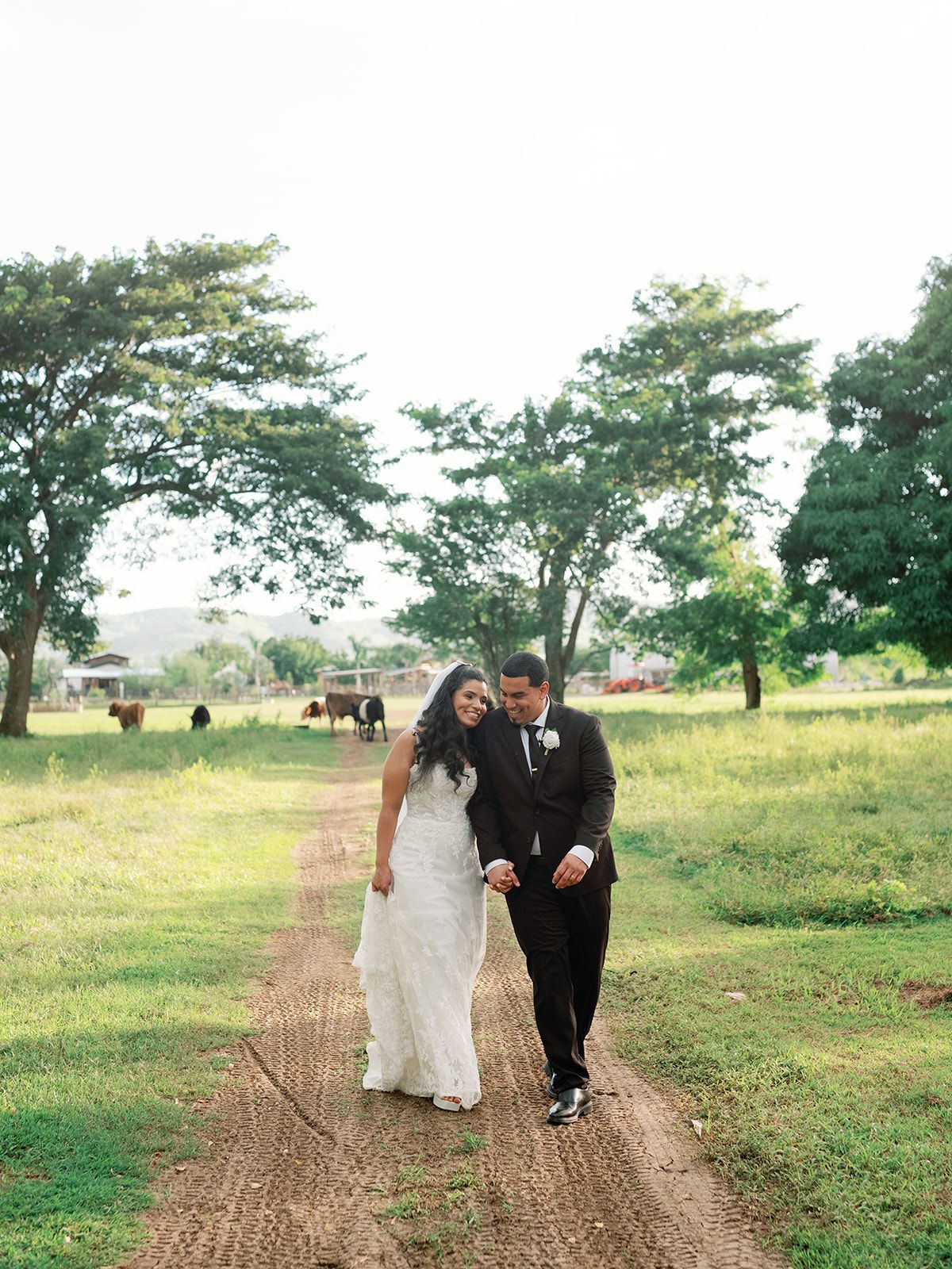 A couple walking down a dirt path with trees and fields in the background.