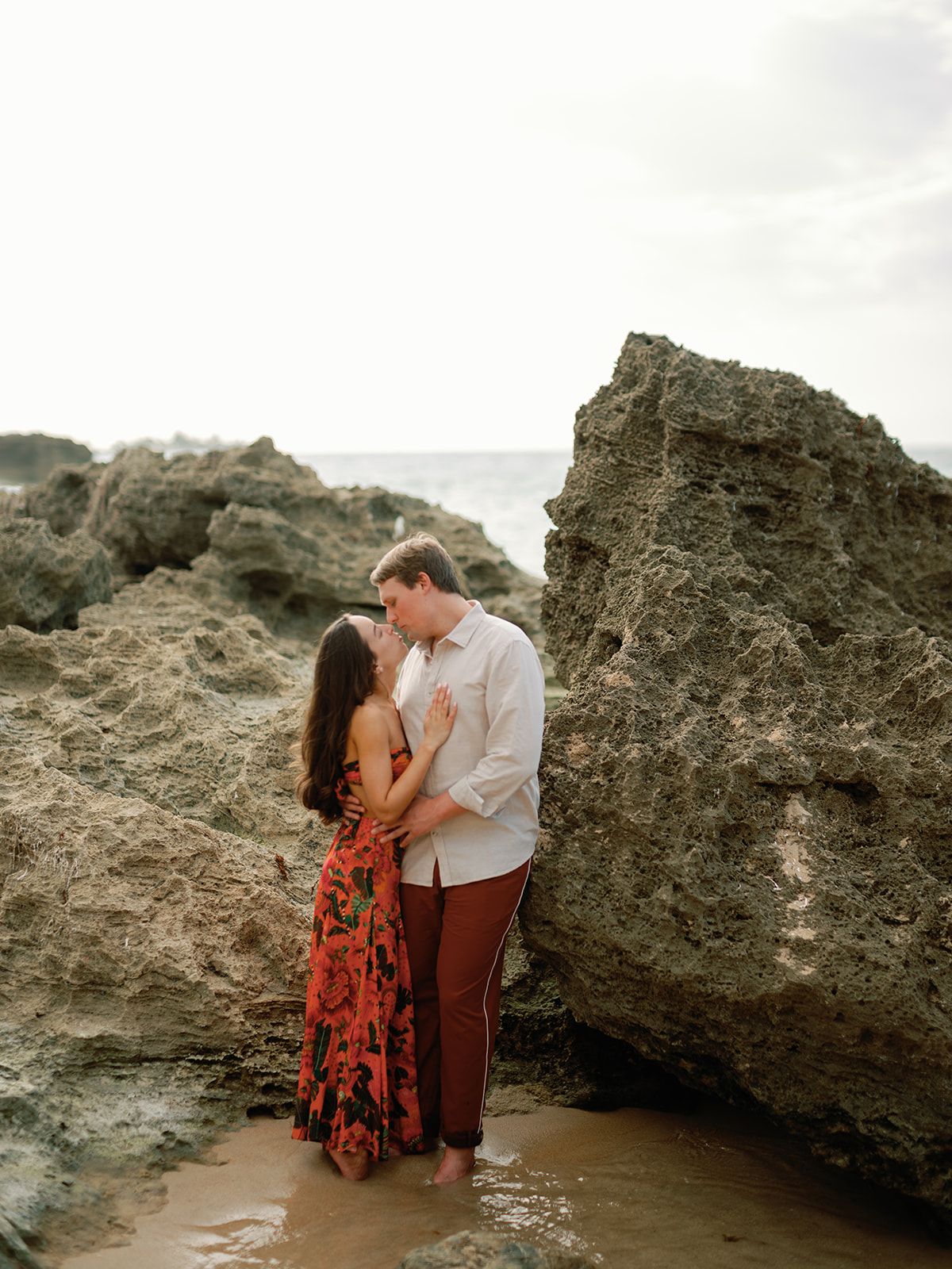 A couple embracing on a rocky beach near the ocean during sunset.