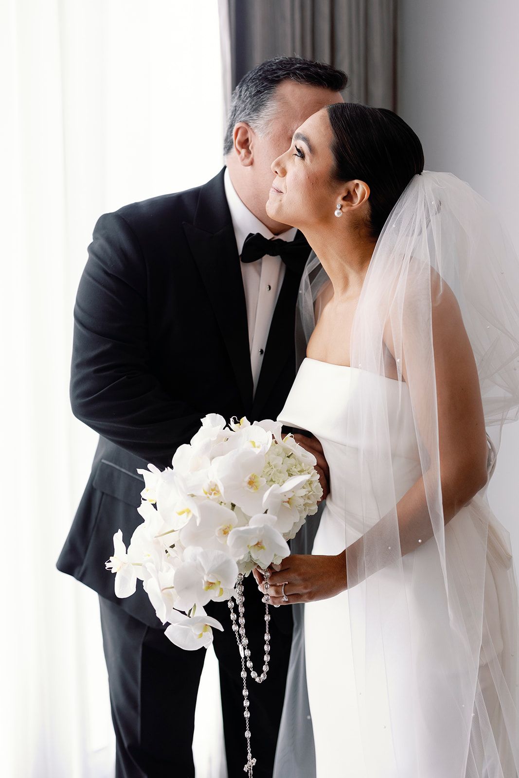 A romantic wedding moment with a beautiful white orchid bouquet, veil, and formal black tuxedo.