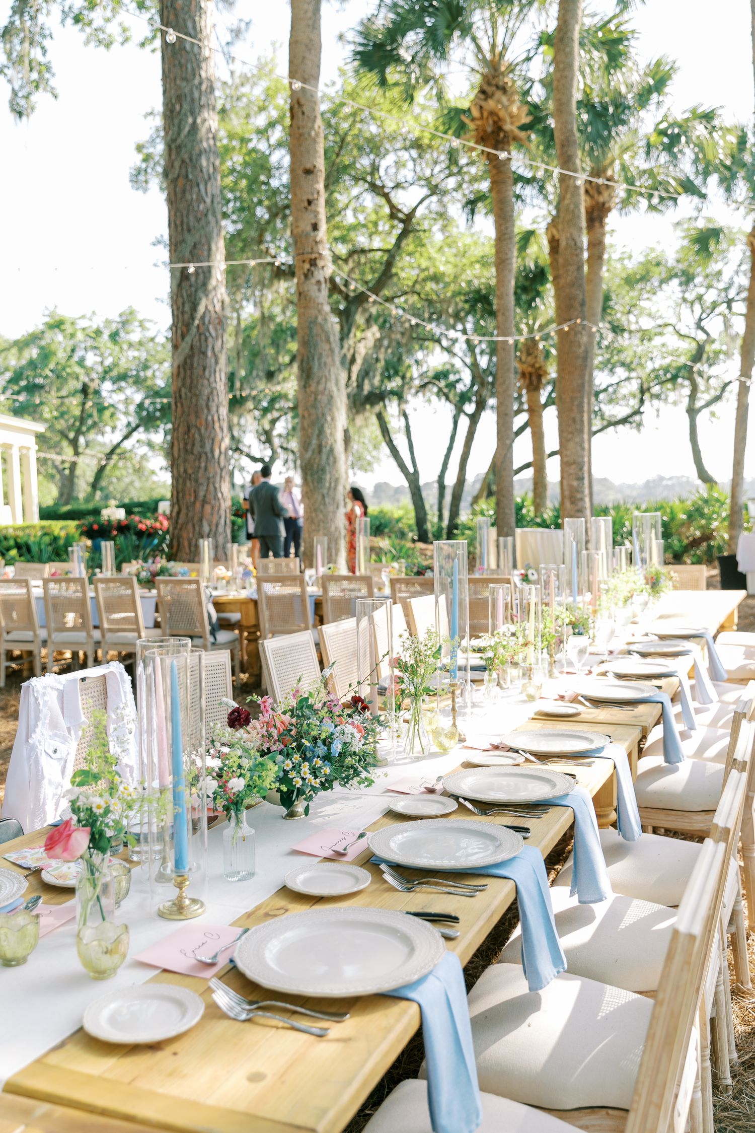 An elegant outdoor wedding reception table setup with white chairs under tall pine trees.