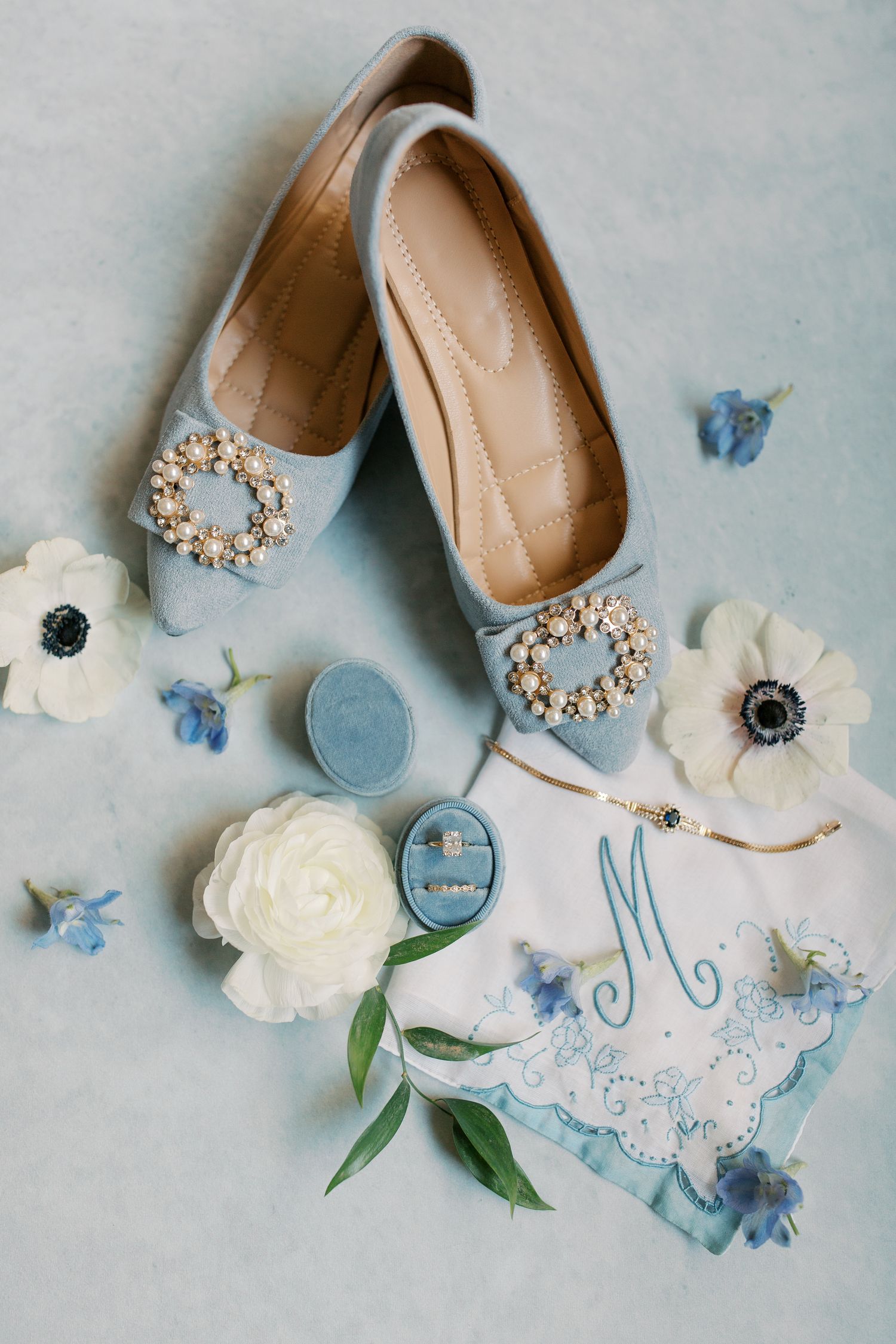 Wedding accessories including bridal shoes, blue details, and white flowers arranged on a light blue surface.