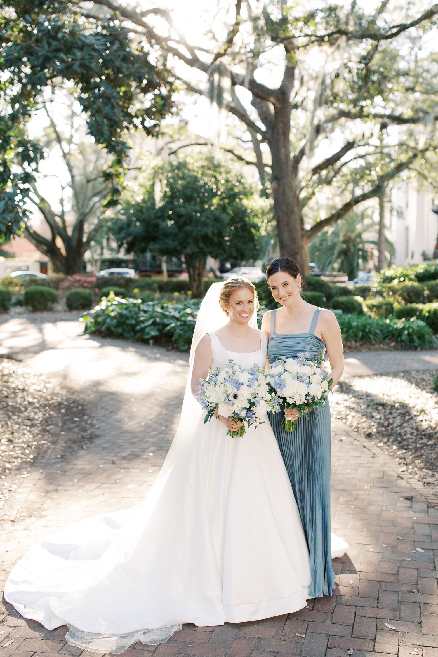 Wedding party members in white and blue dresses walk along a brick pathway under oak trees.
