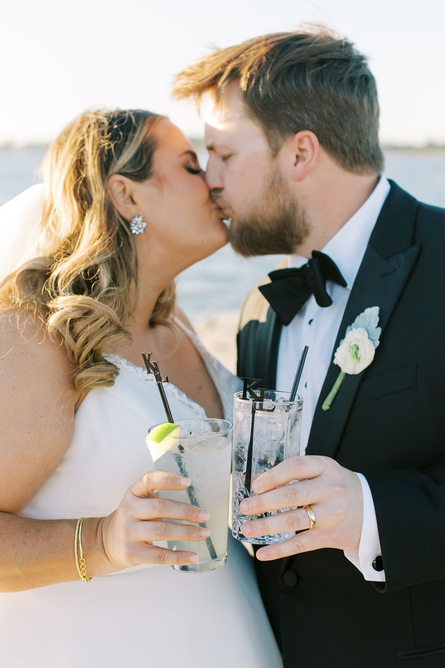 Newlyweds share an intimate kiss while holding cocktails during a sunset beach wedding celebration.