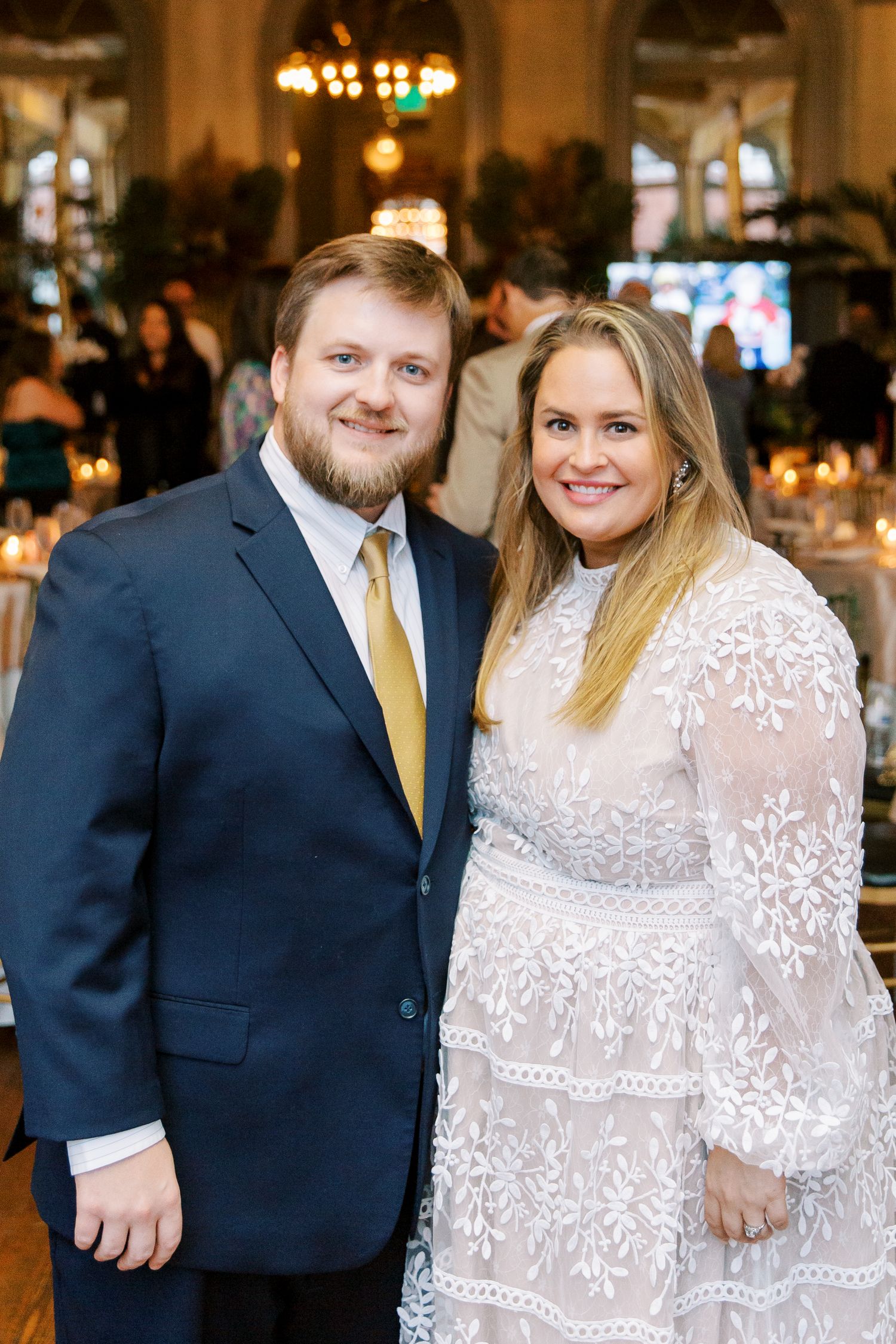 A couple poses together at an elegant indoor venue, with the woman wearing a white lace dress and the man in a navy suit.
