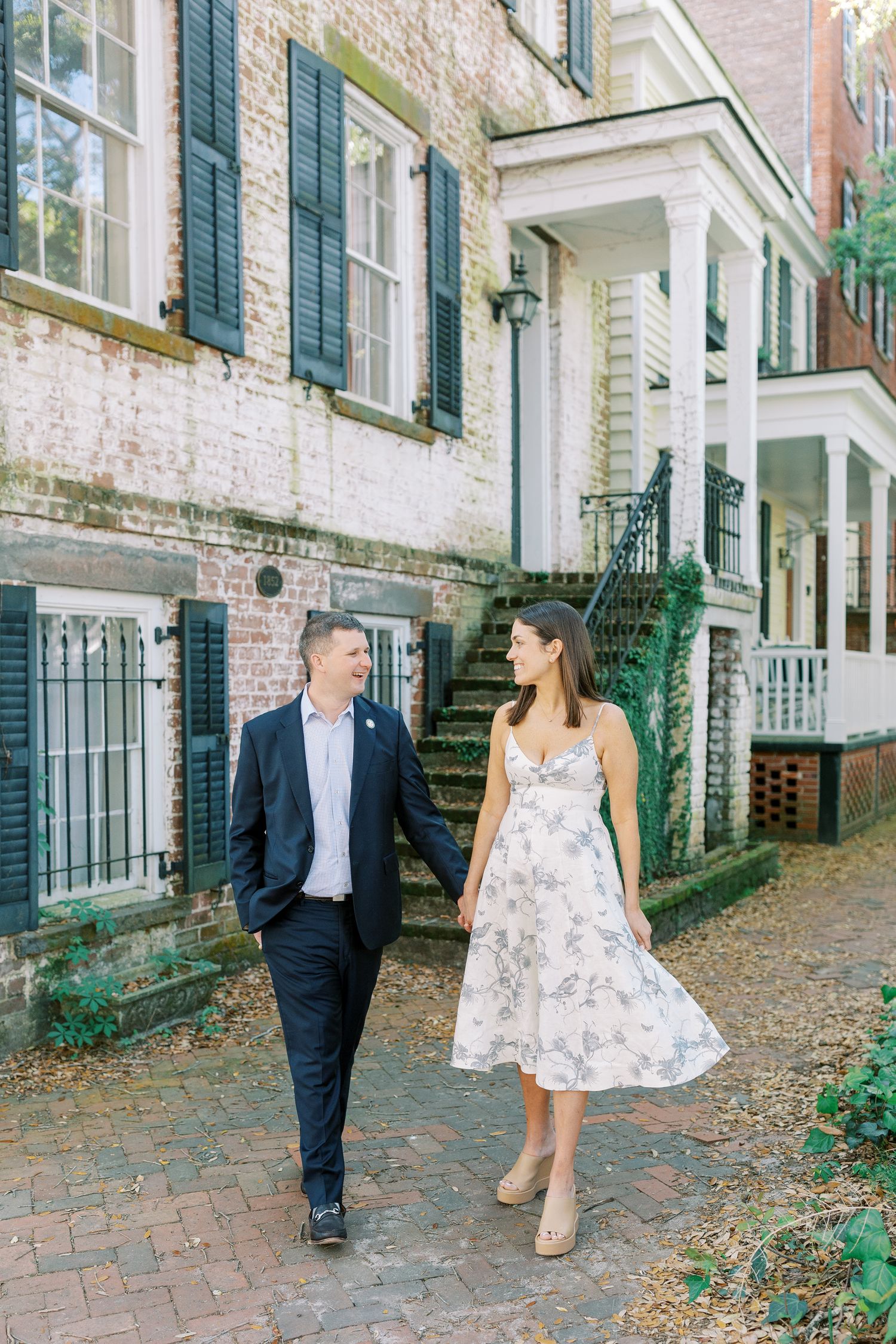 A couple strolling together on a historic brick sidewalk in front of elegant townhouses with green shutters and white columns.