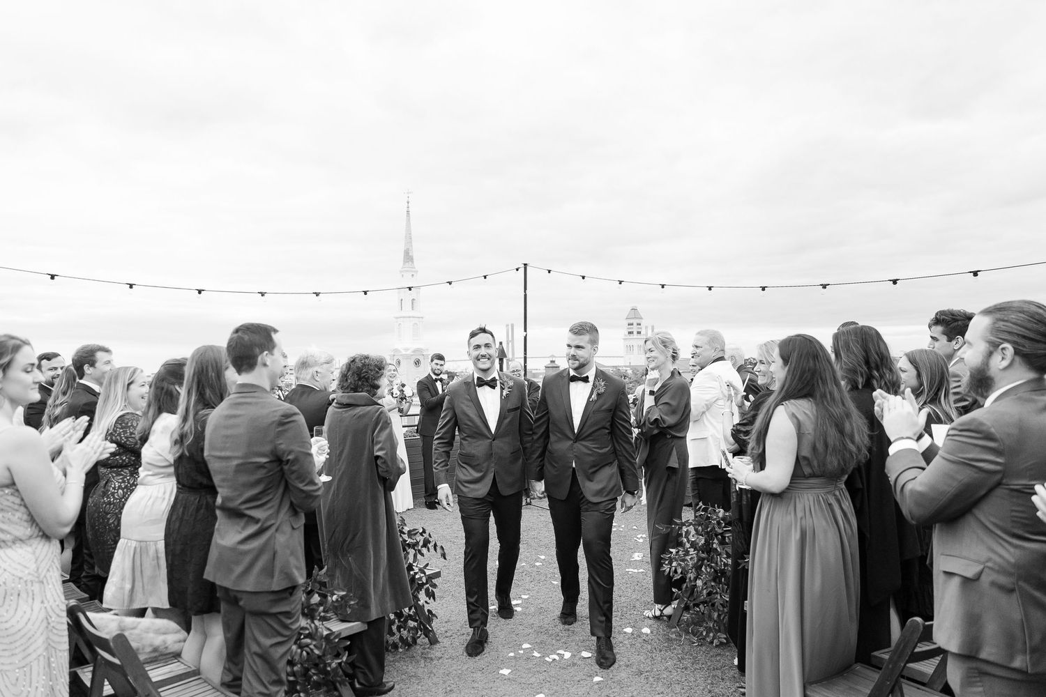 Wedding party gathered outdoors on the rooftop of Perry Lane with string lights above in black and white photo.