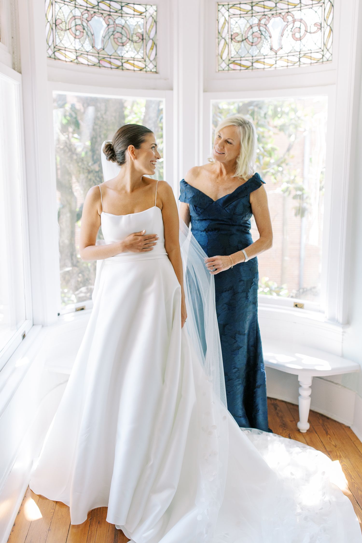 Two women in elegant dresses share a moment in a sunlit room with stained glass windows.