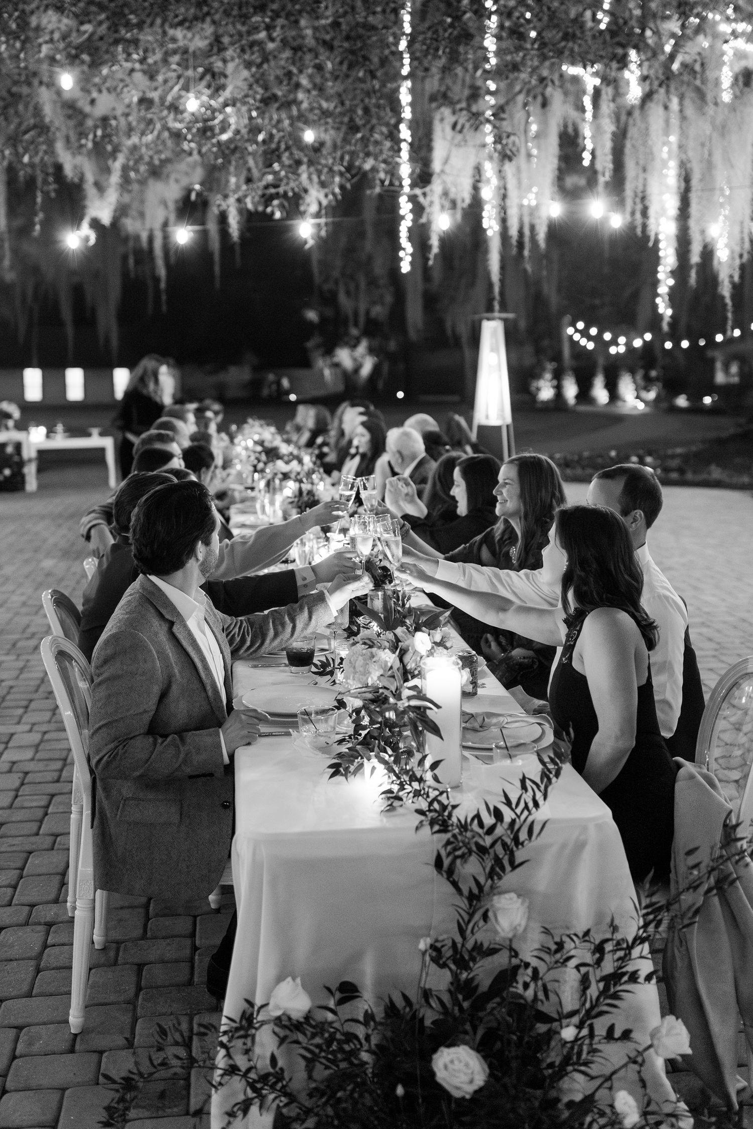 An elegant outdoor wedding reception under hanging lights with guests toasting at a decorated table at Hewitt Oaks in Bluffton.