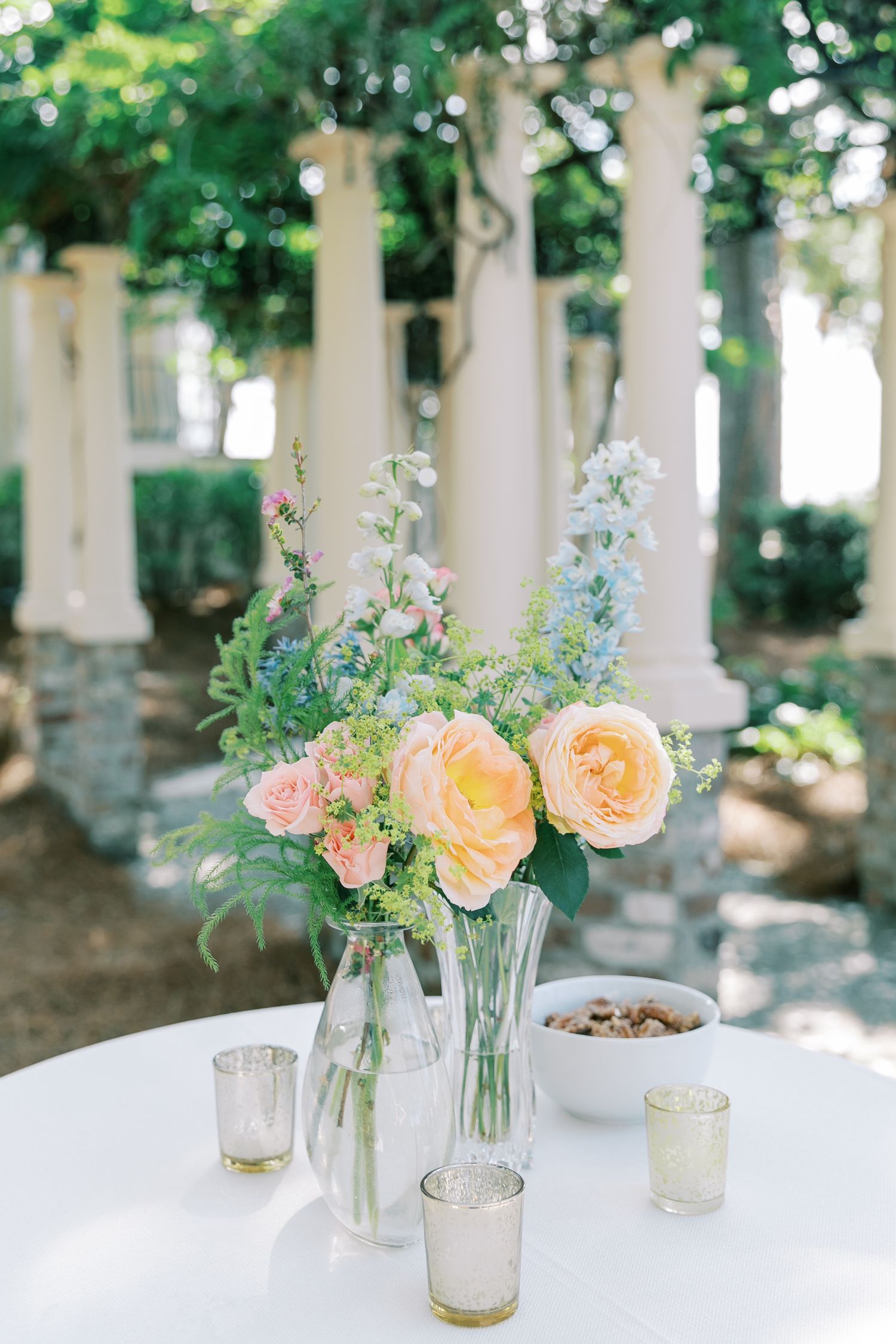 Elegant floral centerpiece with peach roses and blue flowers in glass vases on white table.