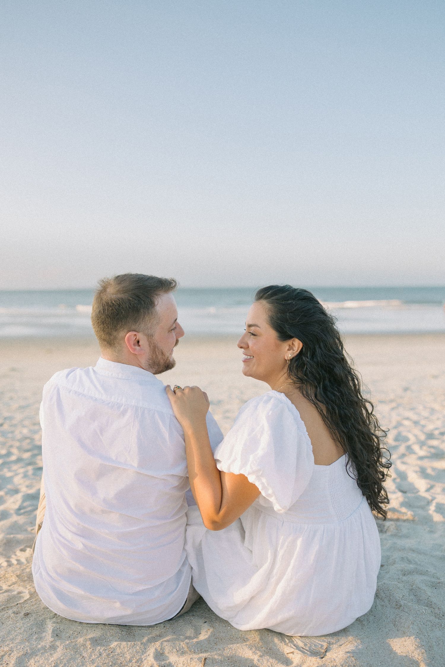 Couple in white outfits sitting close together on a sunny beach.