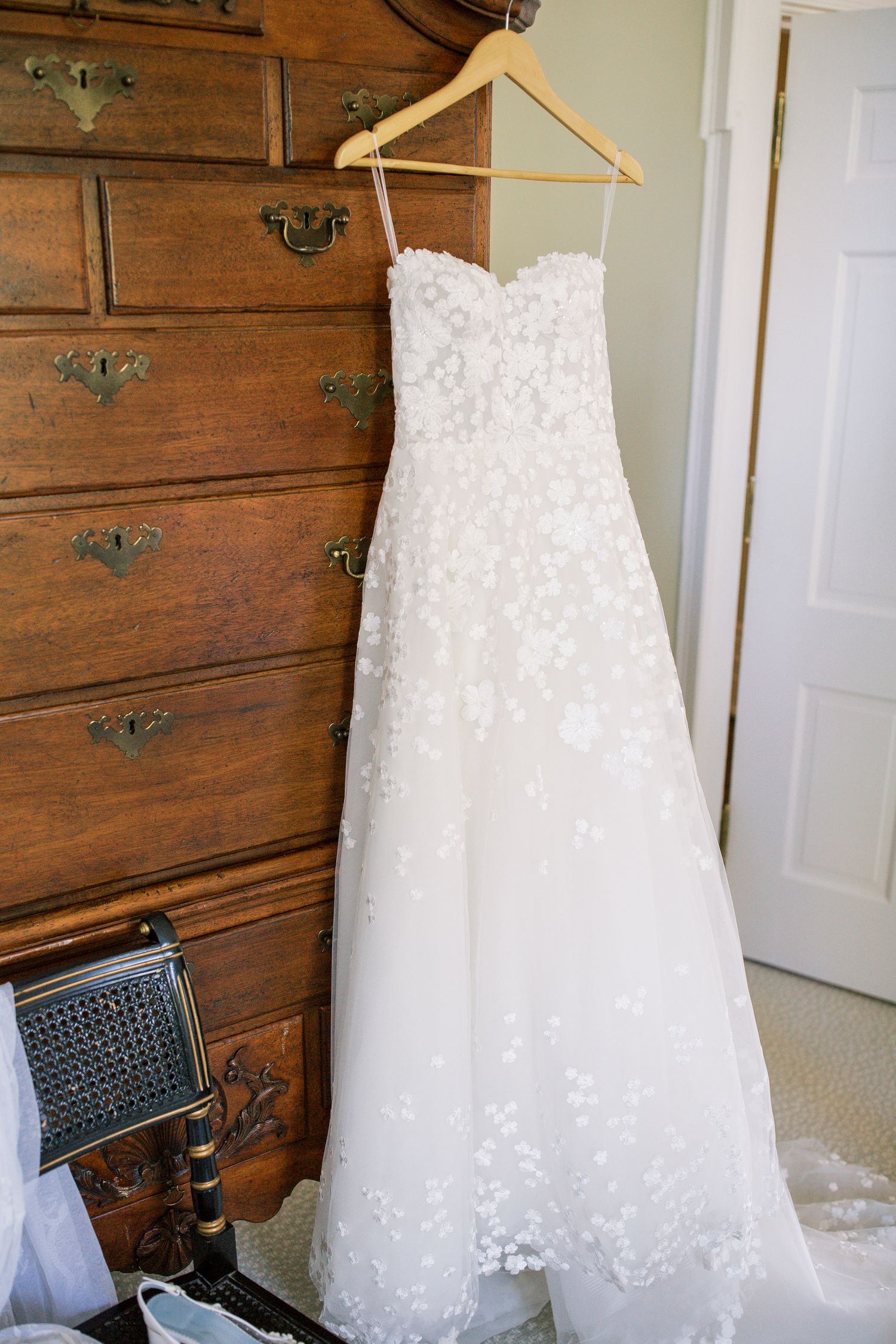 White strapless wedding dress with lace details hangs on wooden hanger against dark wooden furniture background.