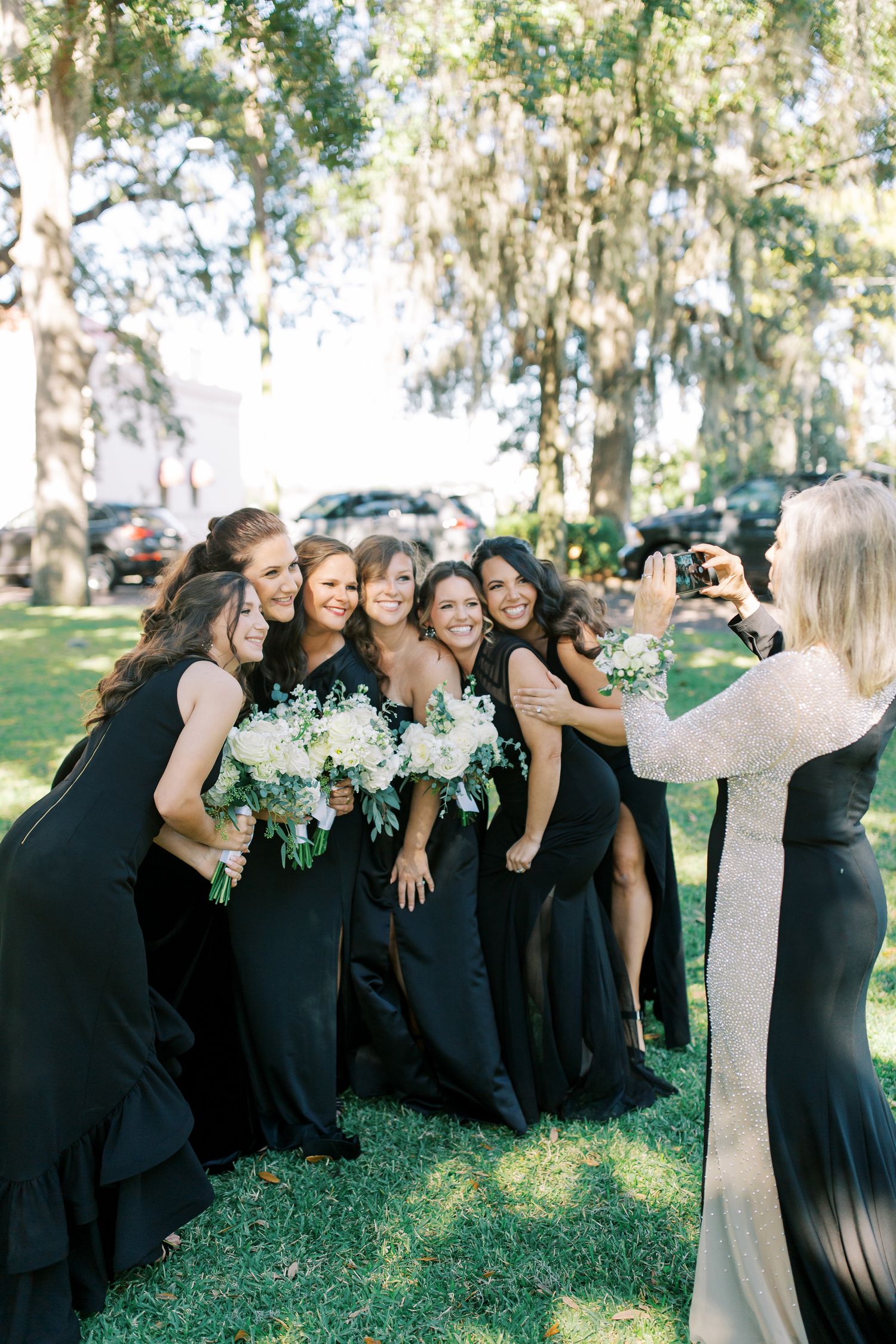 A group wearing matching black gowns pose together on a sunny lawn with Spanish moss trees in the background.