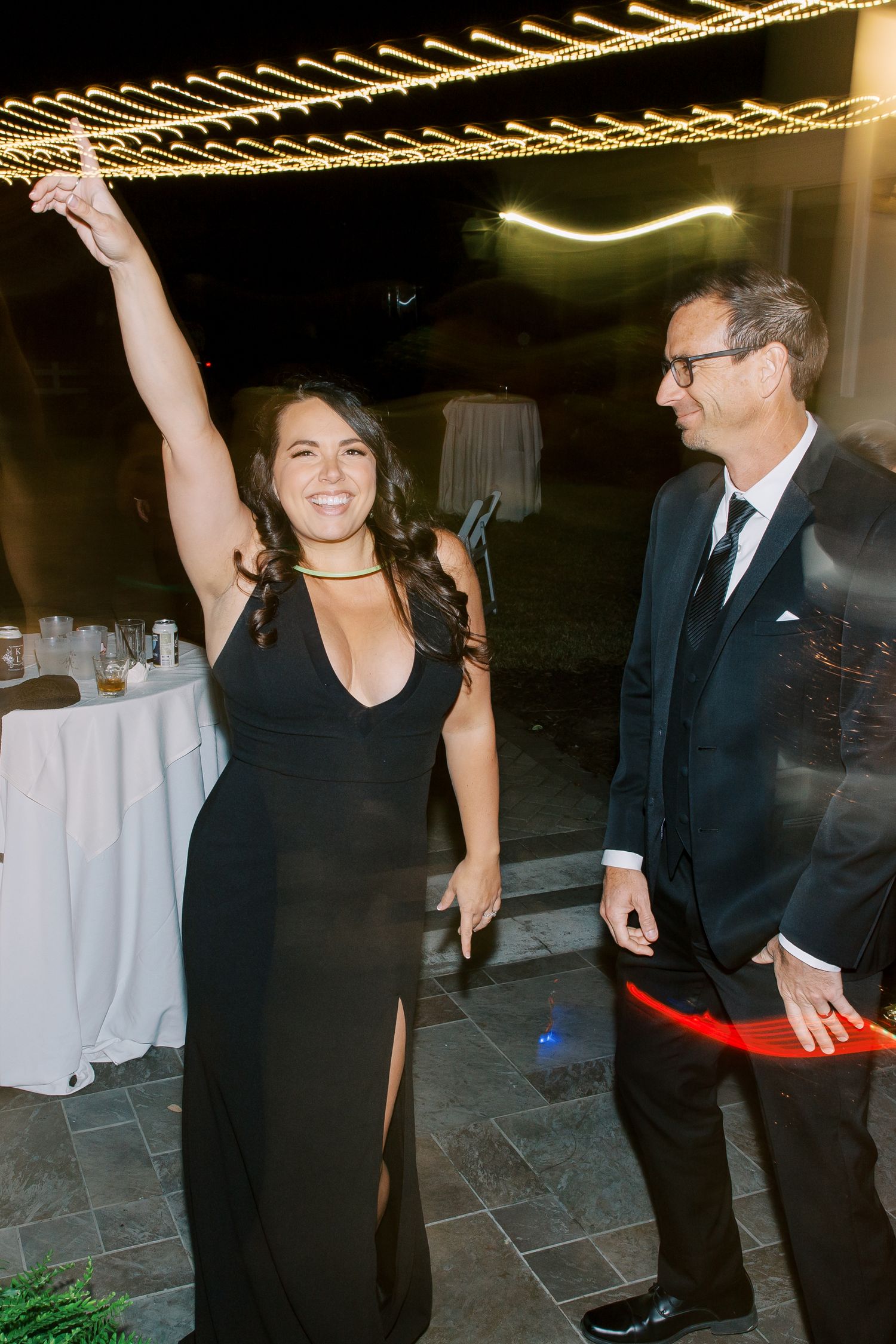 Two wedding guests enjoying a celebratory dance moment under string lights at an evening reception.