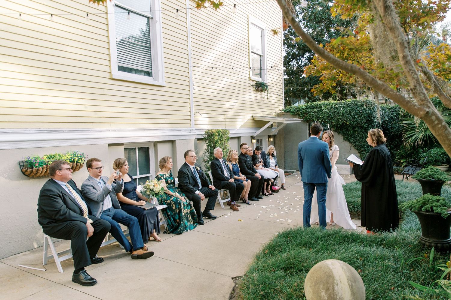 Wedding guests seated along a cream-colored building exterior during an outdoor ceremony.