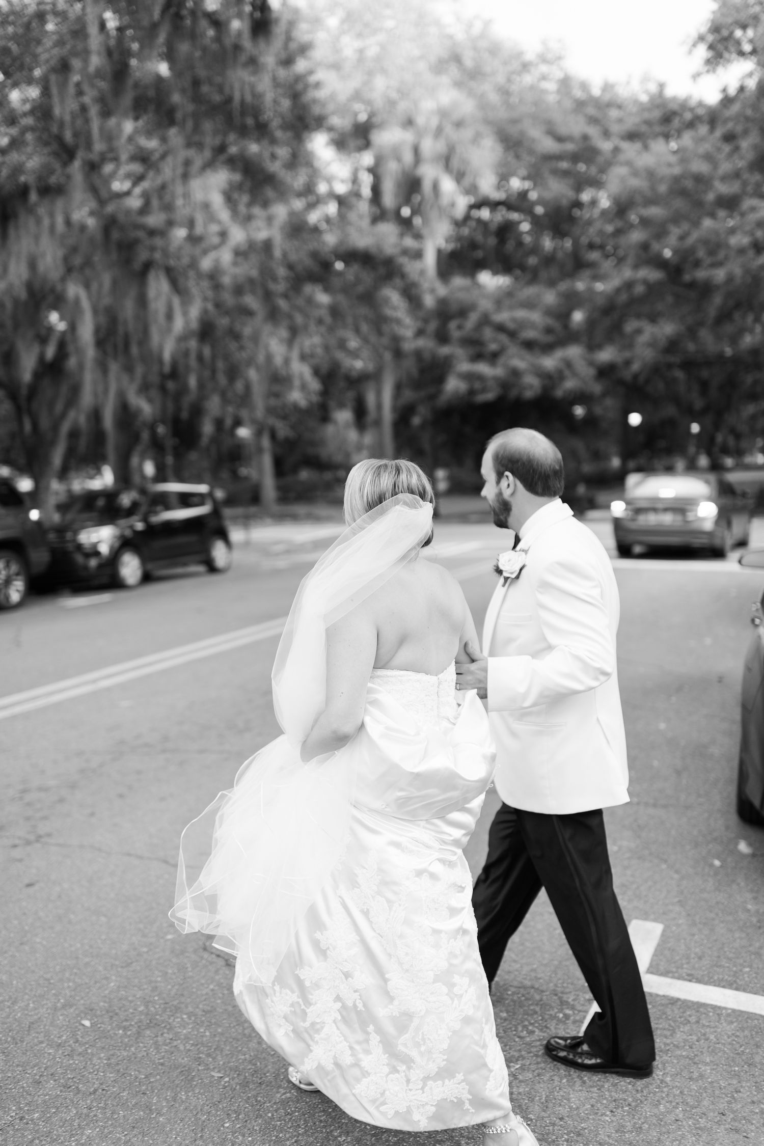 Black and white photo of couple walking together after wedding ceremony on tree-lined street.