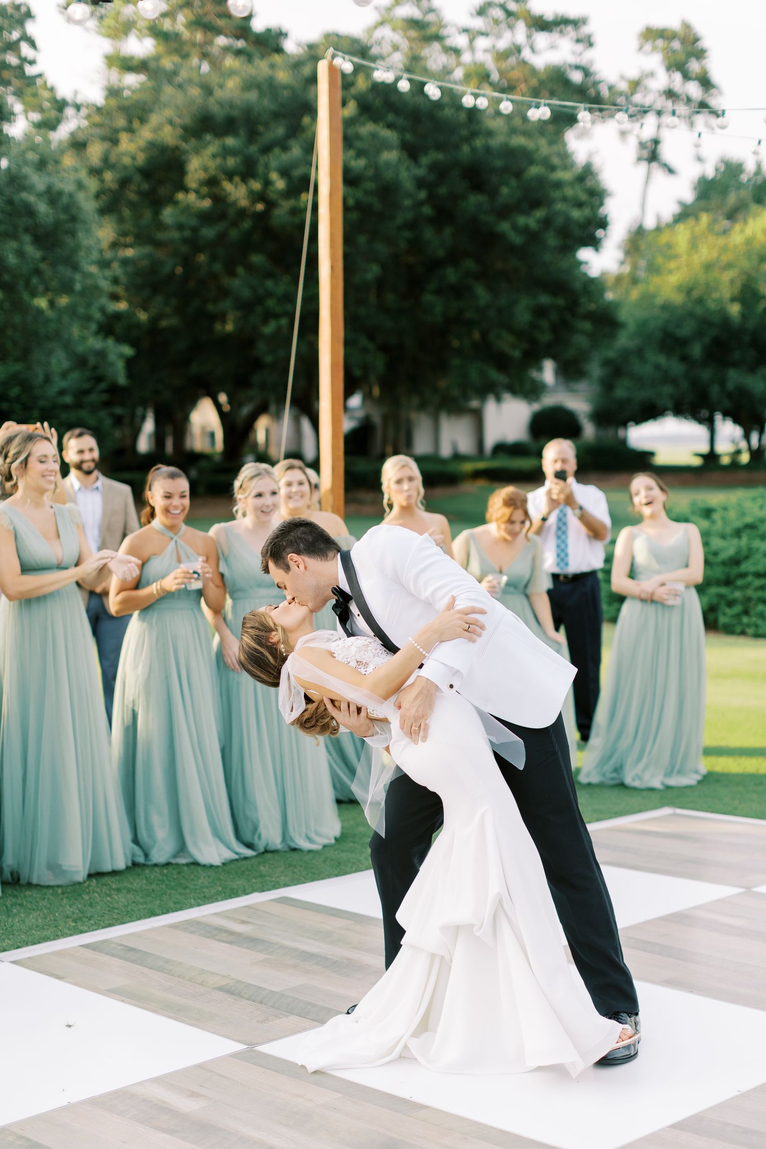 Elegant couple performs romantic dance dip on checkerboard dance floor surrounded by bridesmaids in blue dresses.