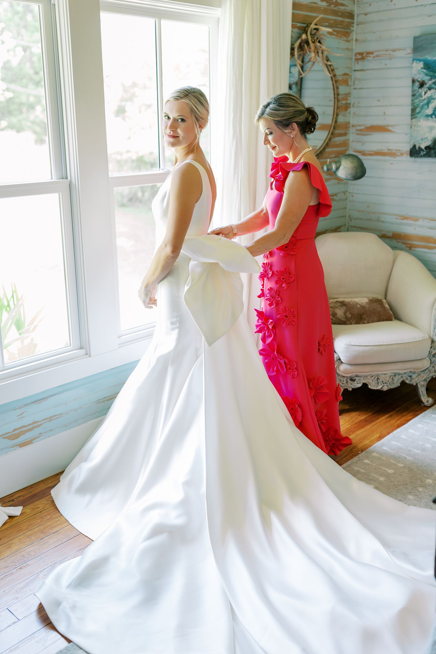 Elegant white wedding dress being fitted on in a bright sunlit room with blue coastal decor.