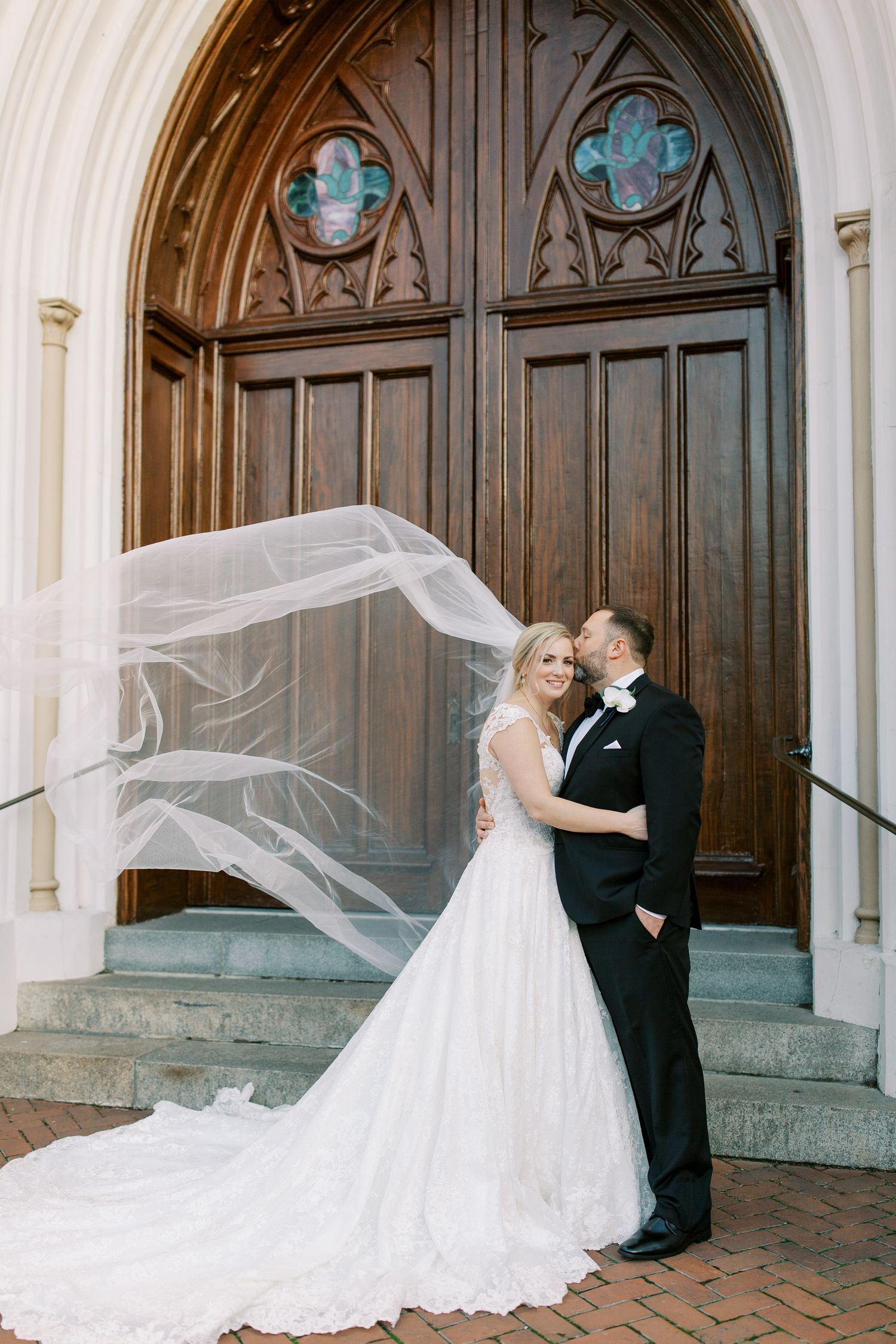 Romantic wedding portrait in front of grand wooden church doors with flowing veil in breeze.