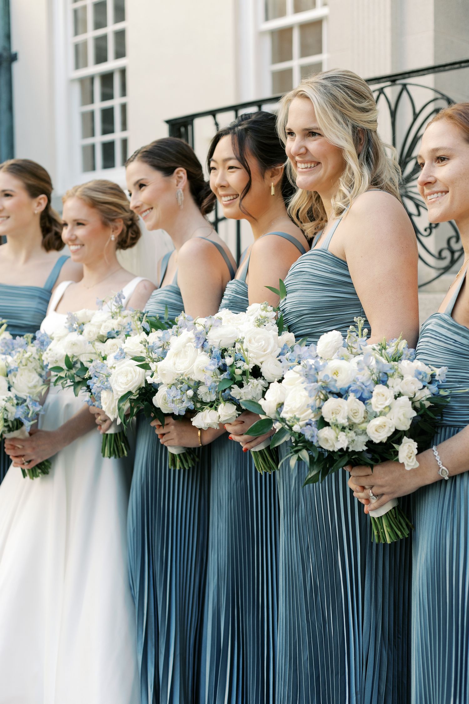 Bridesmaids in blue pleated dresses hold white and blue floral bouquets while standing in a row.