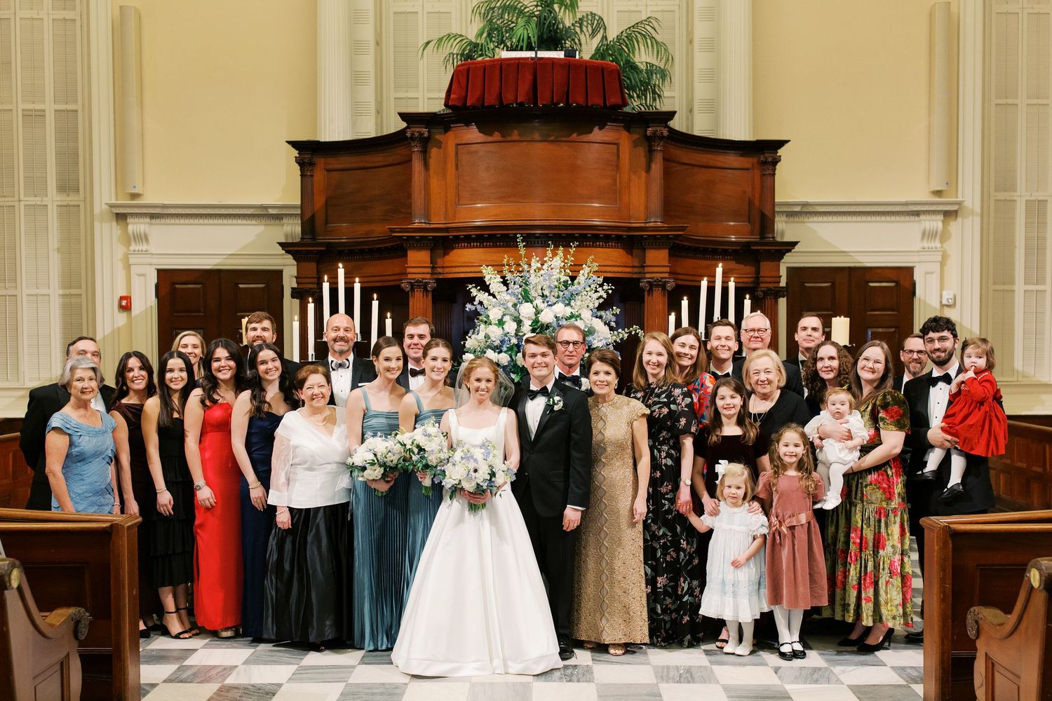 Large wedding party gathered in church sanctuary with ornate wooden altar and formal attire.