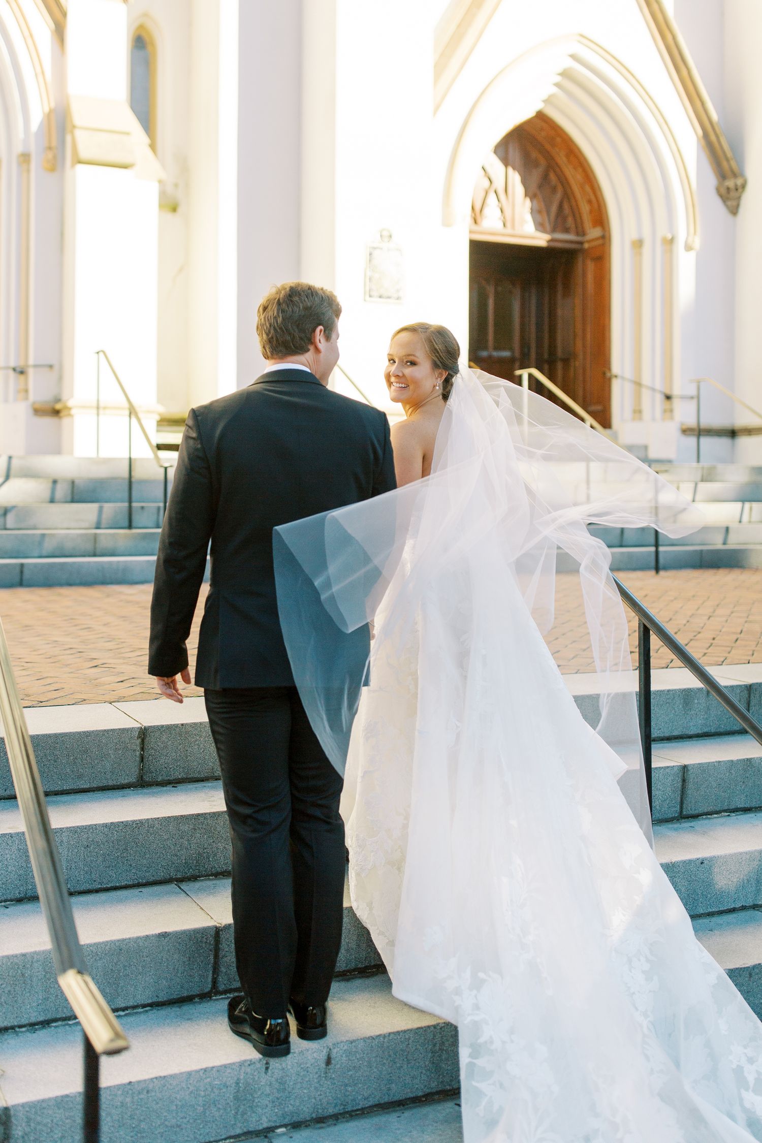 Wedding couple ascending church steps with flowing veil trailing behind.