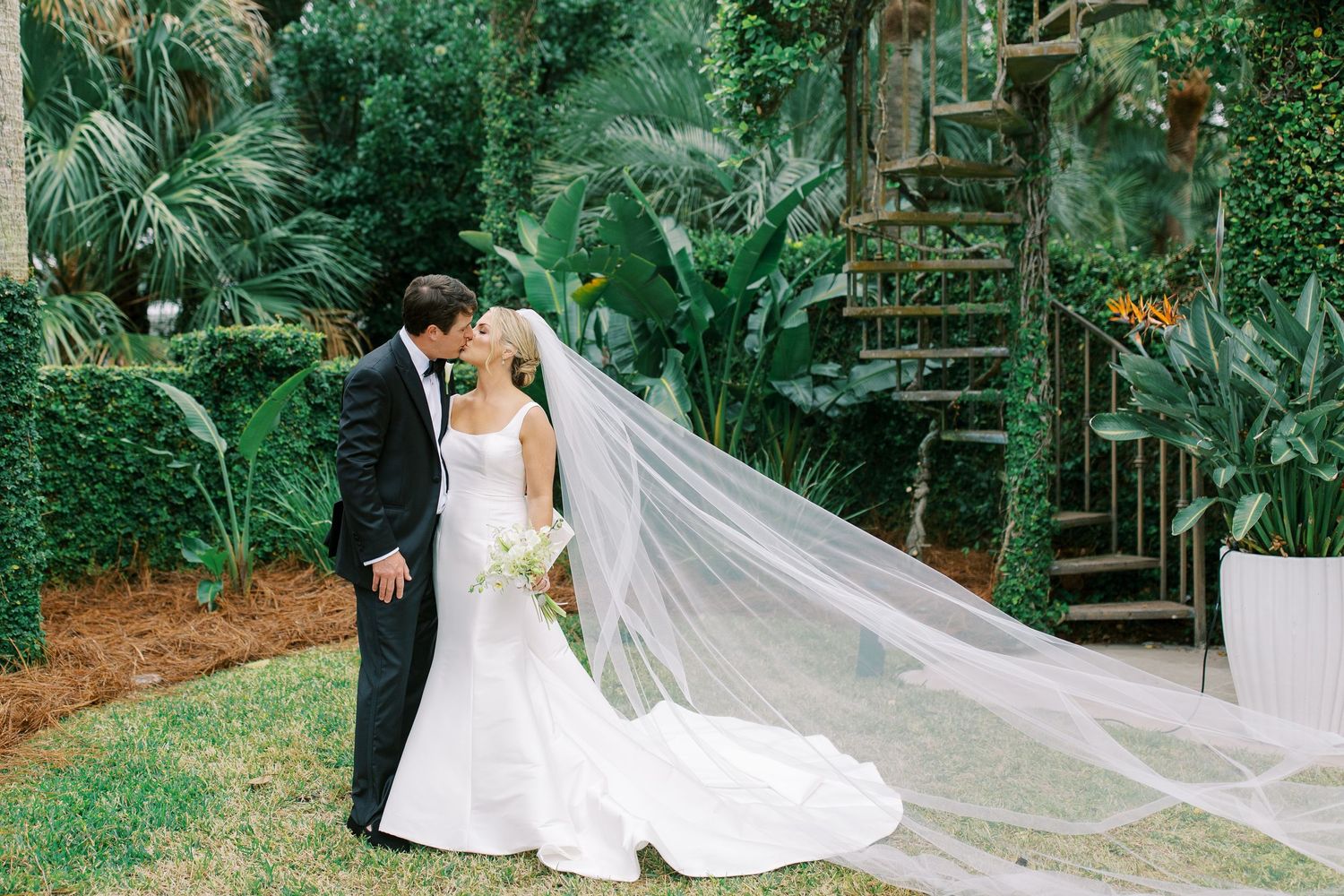 A wedding couple shares an intimate moment in a tropical garden setting with flowing veil.