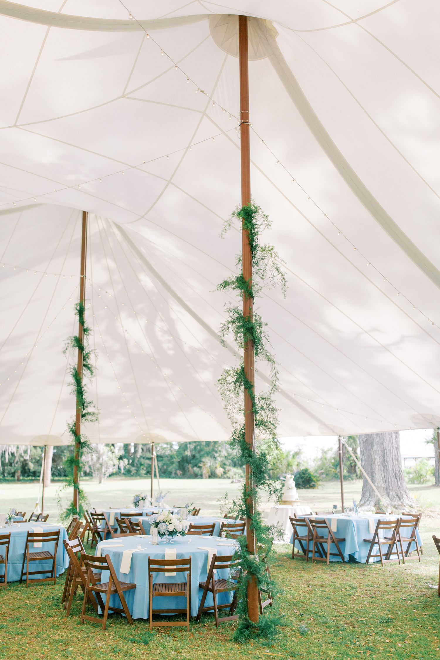 White wedding tent setup with greenery-wrapped poles and round tables with blue linens.