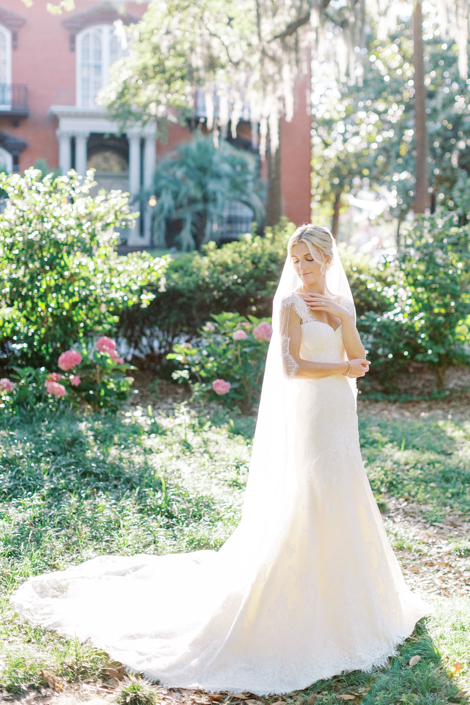 A bride in a flowing white wedding gown with long veil stands in a sunlit garden with historic architecture backdrop.