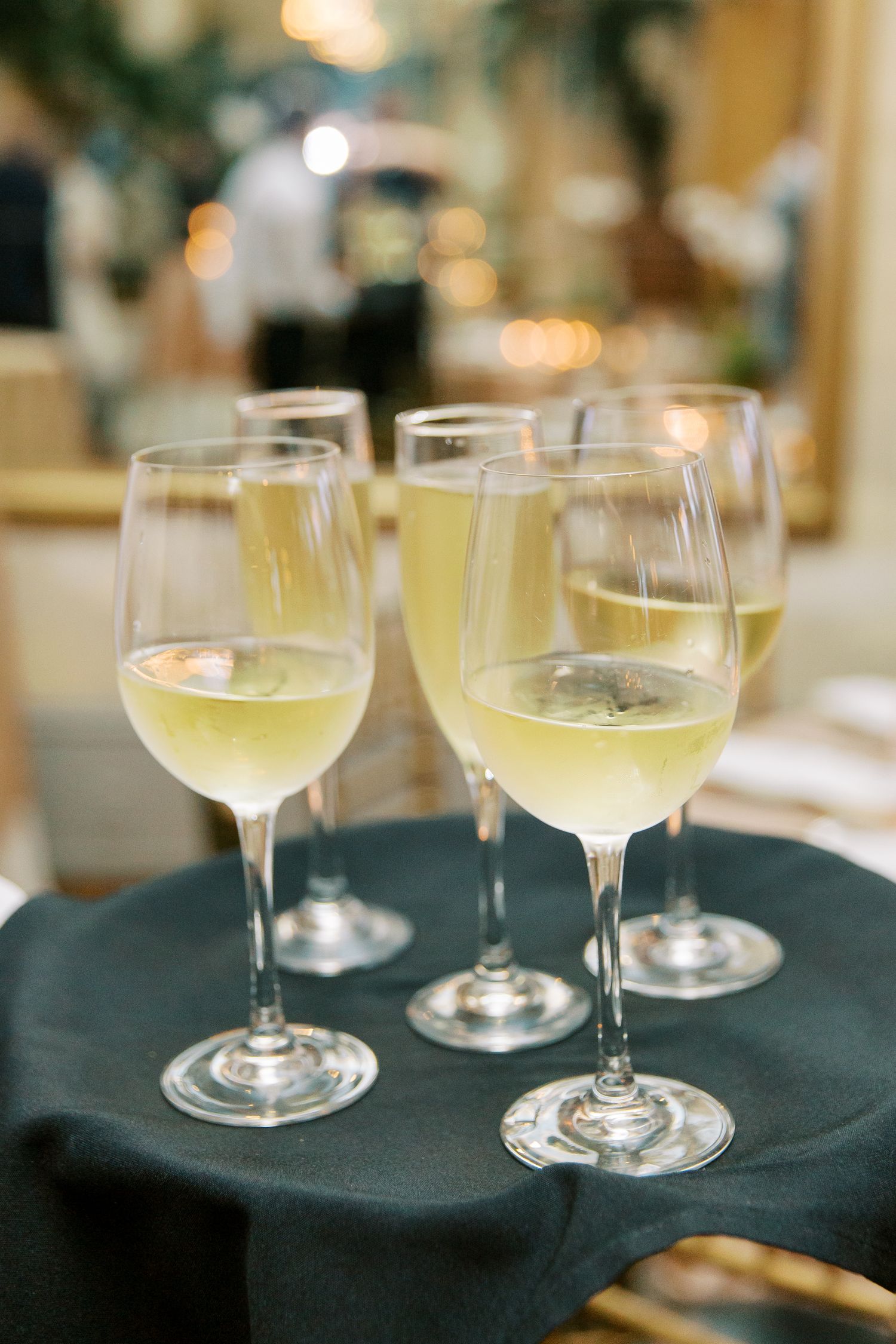 Four elegant wine glasses filled with chilled white wine on a dark tablecloth with soft bokeh lights in background.