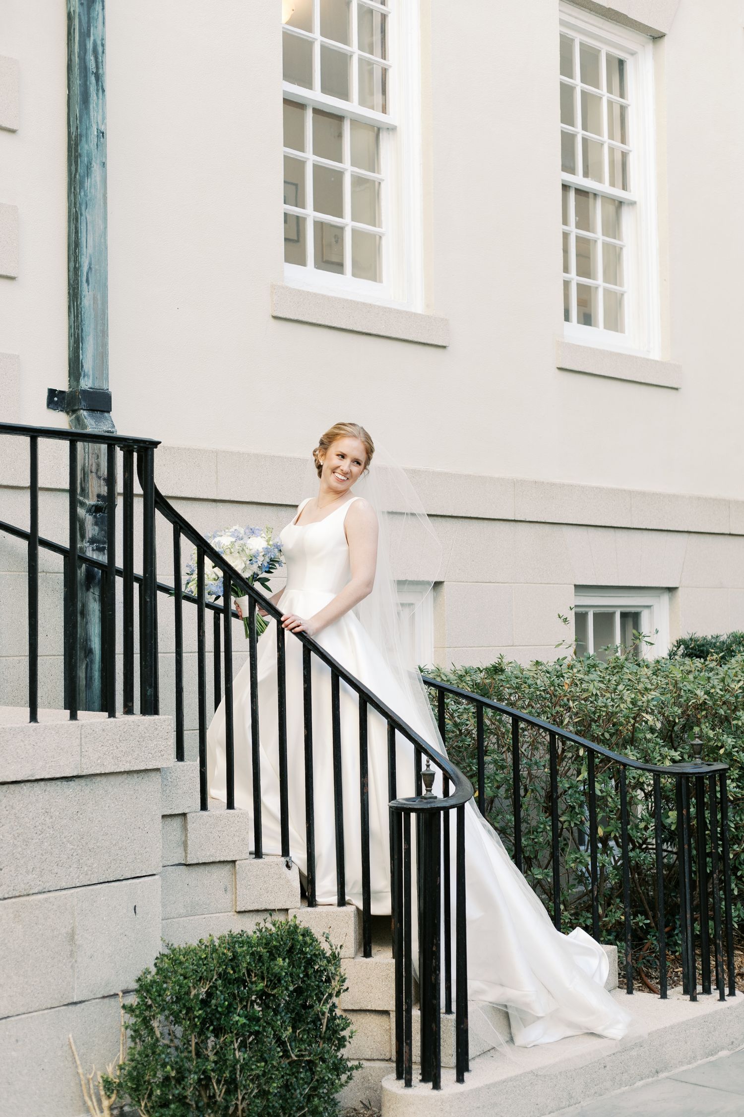 Bride in flowing white gown poses on stone steps with black railing.
