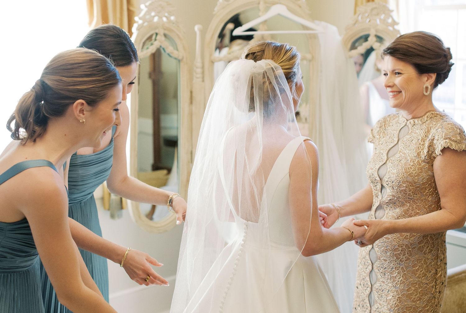 Bridesmaids in blush pink dresses help the bride get ready in an elegant dressing room.