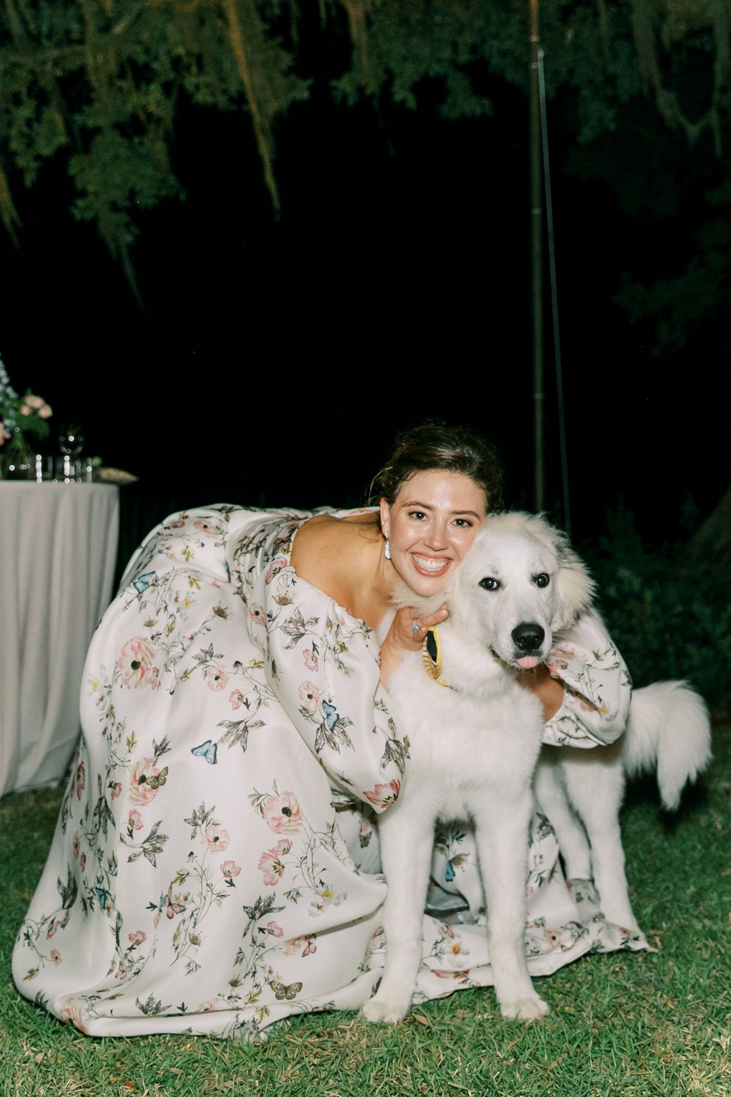 Person in floral dress poses with white dog on grass at nighttime wedding reception.