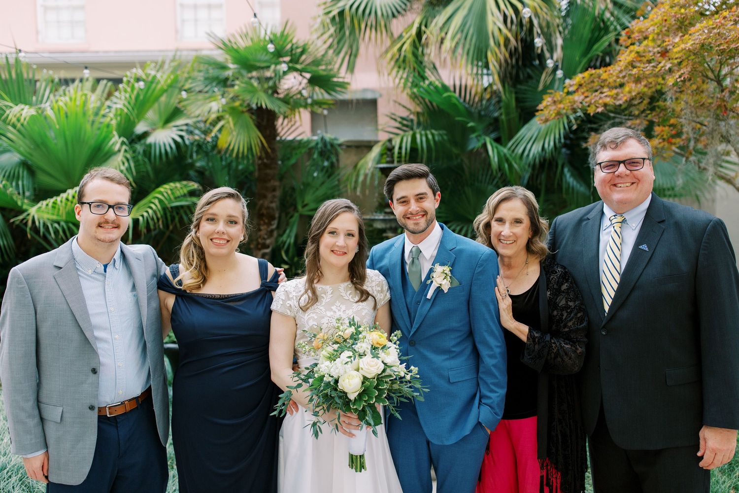 Wedding party in formal attire gathered outdoors with palm trees in the background.
